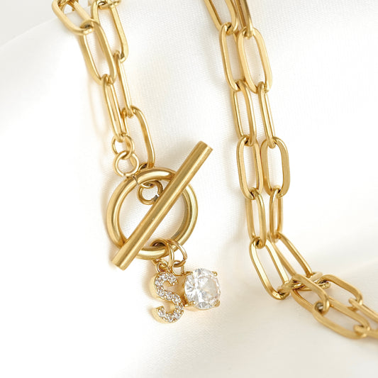 Gold chain necklace with a toggle clasp and a dangling 'S' charm embellished with small clear stones, displayed on a white fabric background.