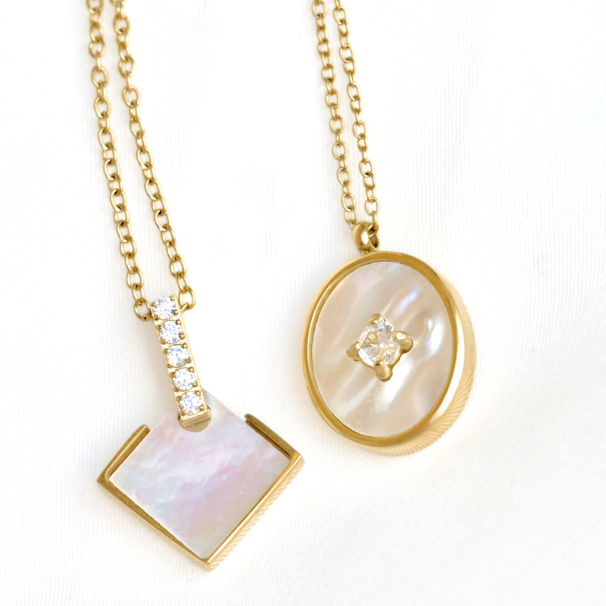 Two gold pendant necklaces with iridescent mother-of-pearl inlays, displayed on a white textured background. One pendant is a square with a bezel-set clear stone, and the other is an oval with a central clear flower-like stone.