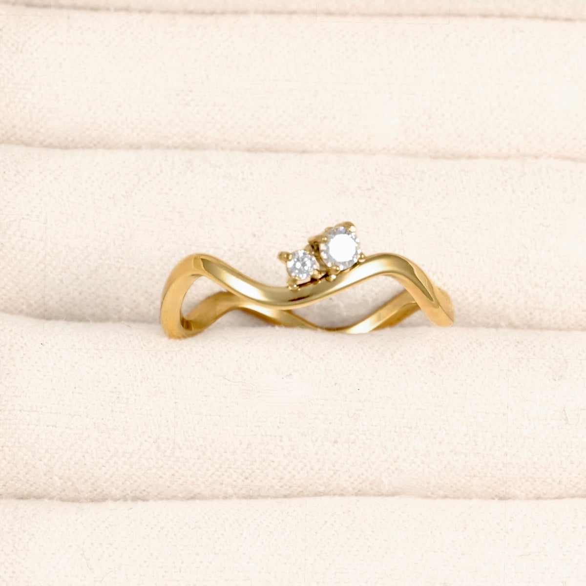A yellow gold ring with a wavy, intertwined band and a small cluster of clear gemstones at the top, displayed on a textured cream-colored ring box.