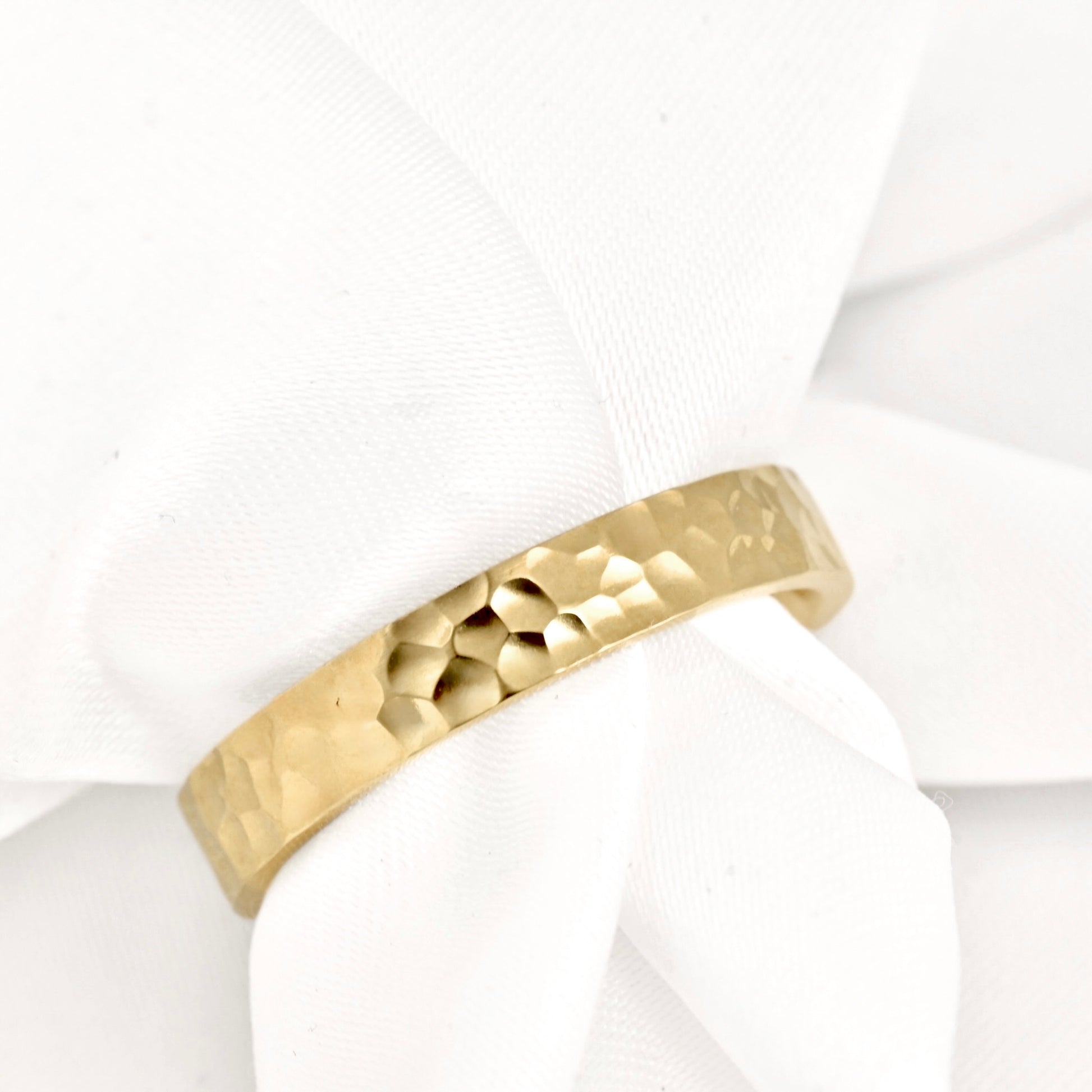 A hammered gold band ring displayed on a soft white fabric ribbon.
