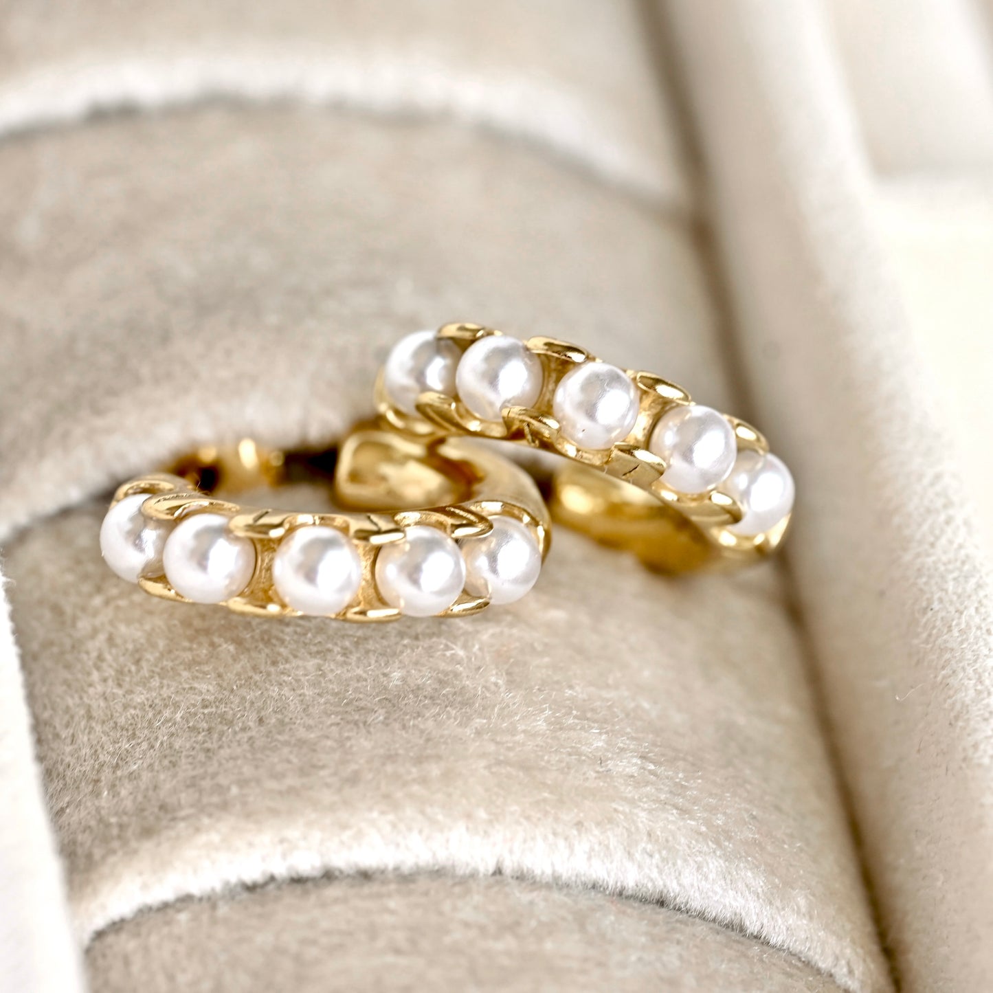 A pair of gold hoop earrings adorned with a row of white freshwater pearls, displayed on a beige velvet jewelry ring holder.