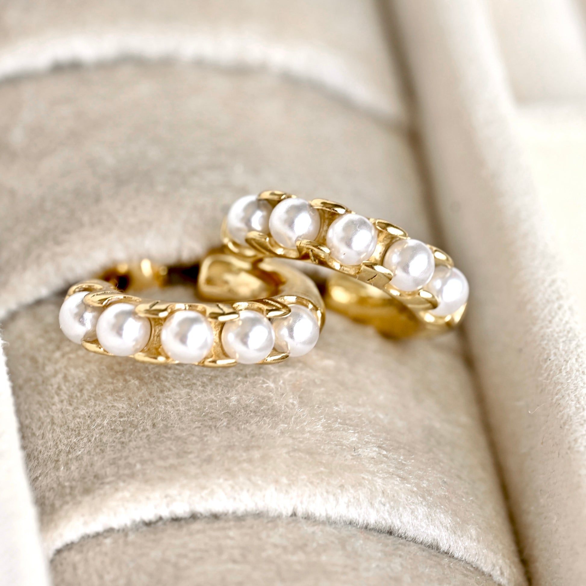 A pair of gold hoop earrings adorned with a row of white freshwater pearls, displayed on a beige velvet jewelry ring holder.