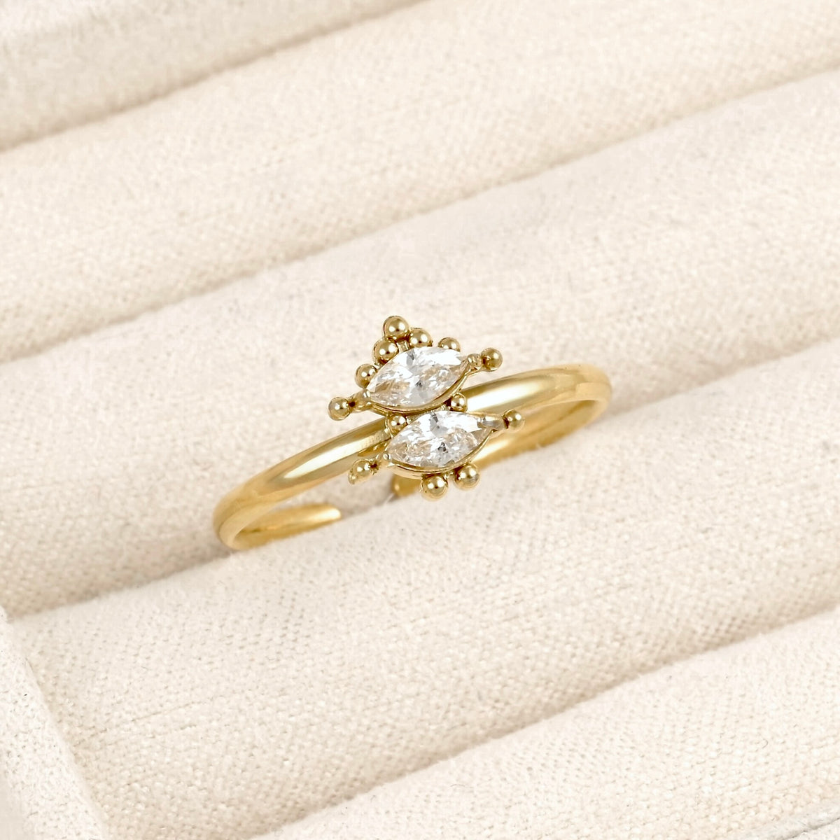 A gold ring with two marquise-cut clear gemstones in a delicate floral cluster setting, displayed on a cream-colored textured fabric ring box.