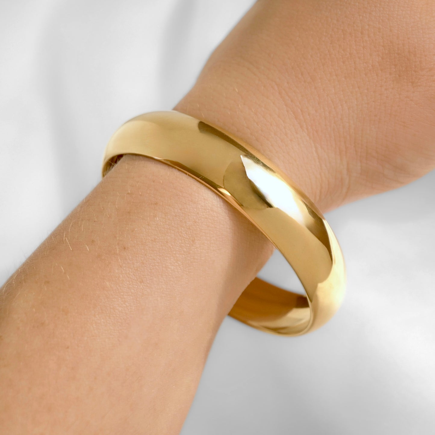 A wide yellow gold bangle bracelet with a dome shape worn on a person's wrist, shown in a close-up shot against a soft white background.