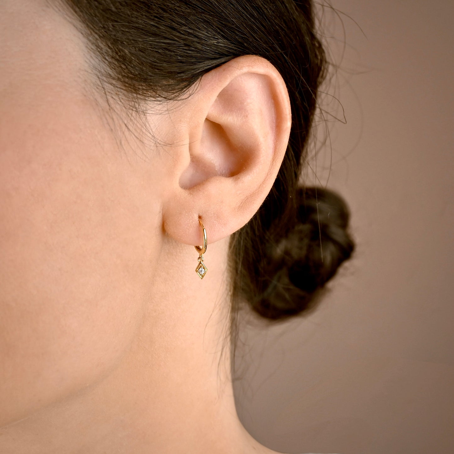 A close-up of a person's ear wearing a small gold huggie hoop earring with a dangling clear gemstone, against a soft beige background.