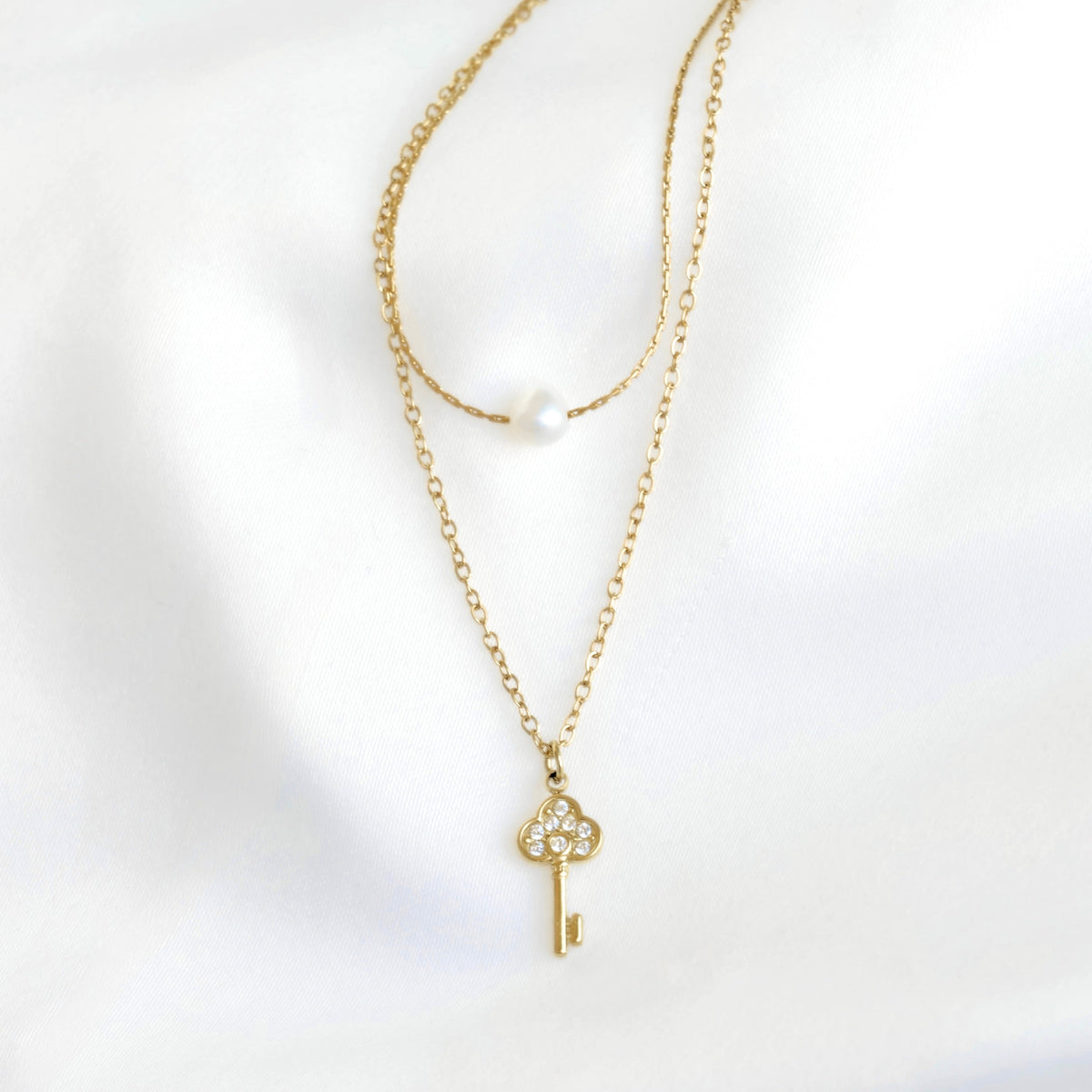 A delicate gold necklace with a key-shaped pendant adorned with small clear stones, displayed on a soft white fabric background. A second layer of chain features a single white pearl bead.