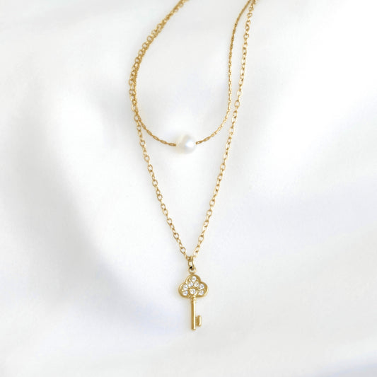 A delicate gold necklace with a key-shaped pendant adorned with small clear stones, displayed on a soft white fabric background. A second layer of chain features a single white pearl bead.