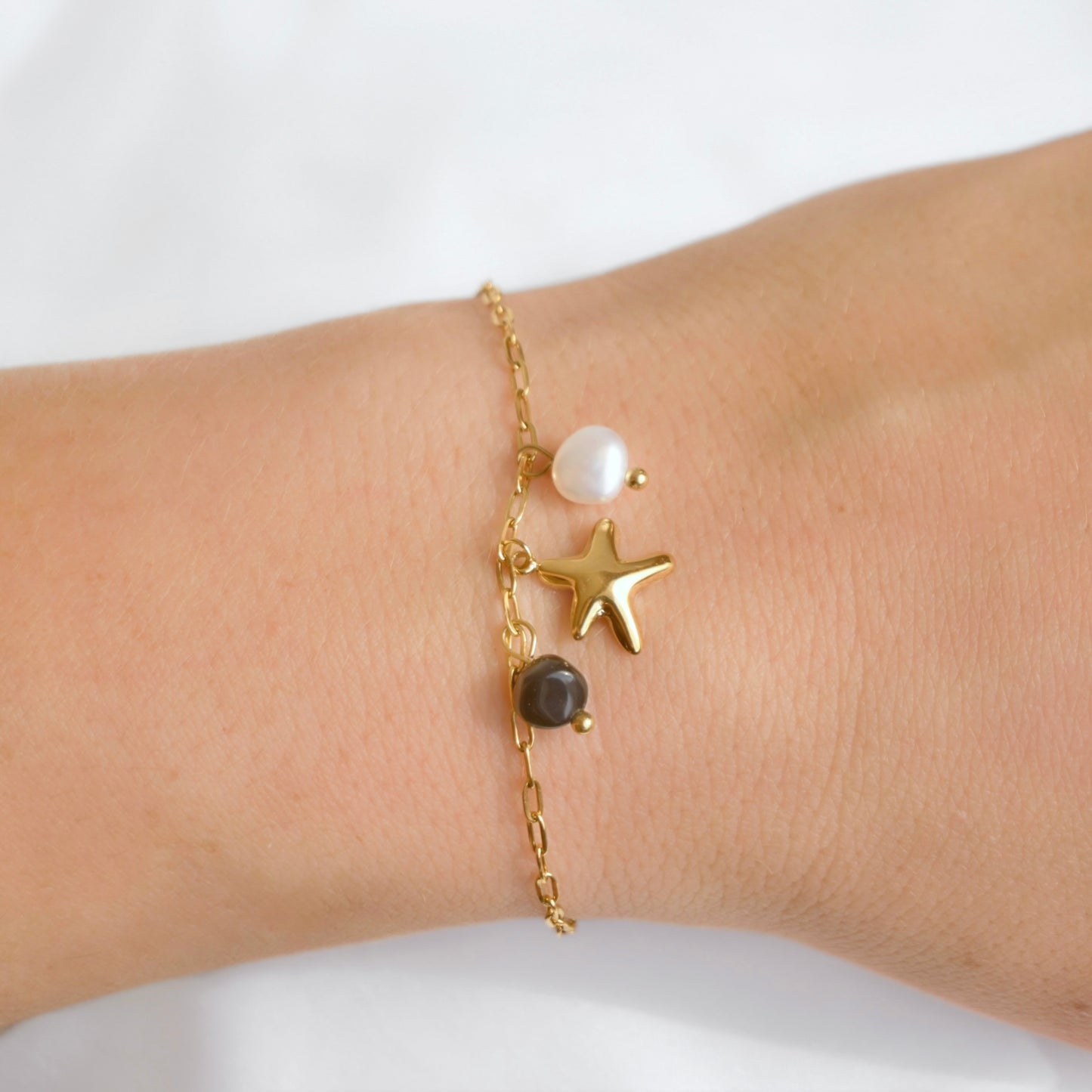 A delicate gold chain bracelet worn on a wrist, featuring a gold starfish charm, a white pearl bead, and a dark gray stone bead.