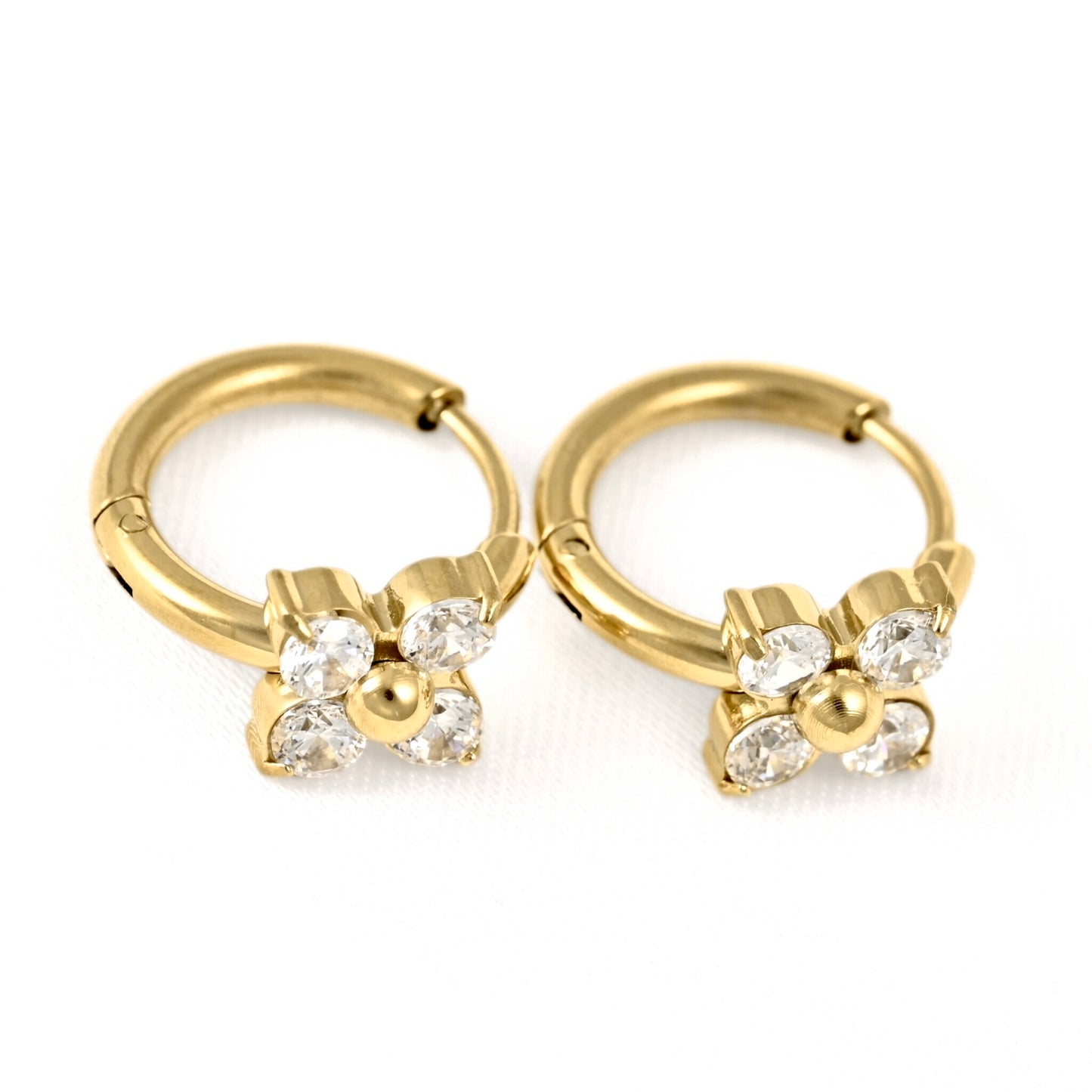 A pair of gold huggie hoop earrings with a small flower charm set with clear rhinestones, shown on a white background.