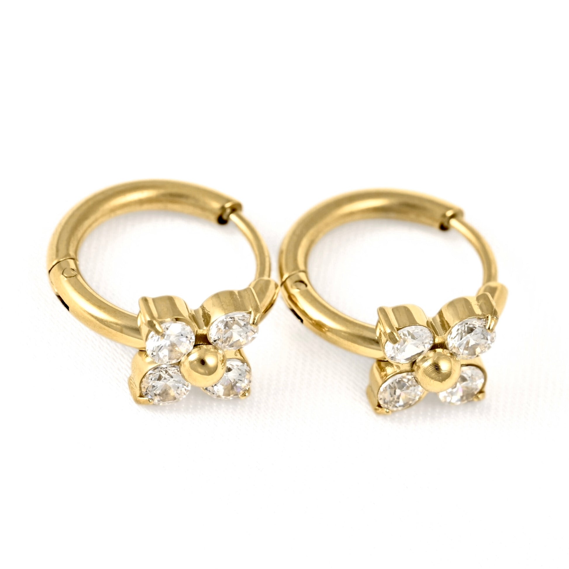 A pair of gold huggie hoop earrings with a small flower charm set with clear rhinestones, shown on a white background.