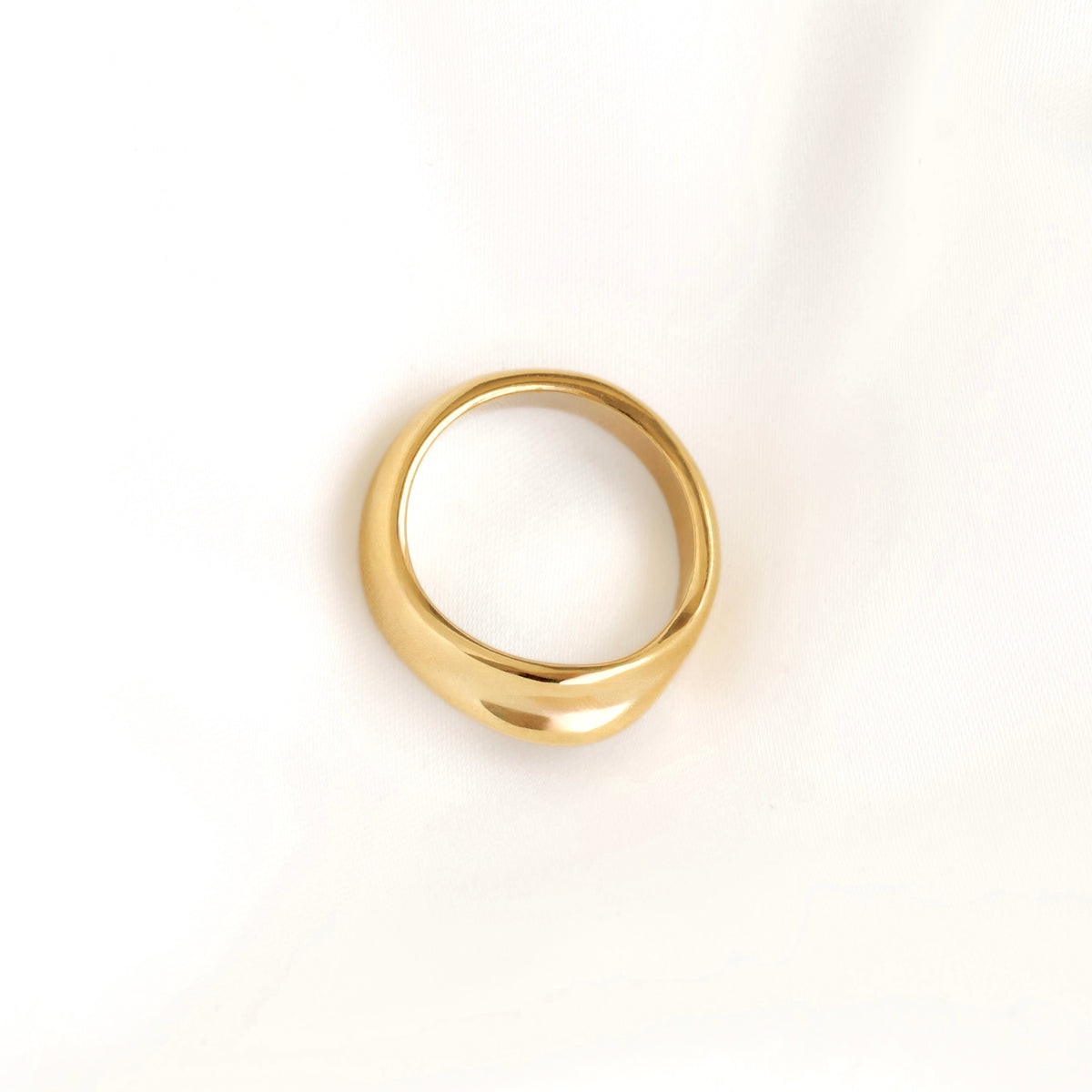 A polished gold signet ring with a wide, rounded band, photographed on a clean white fabric background.