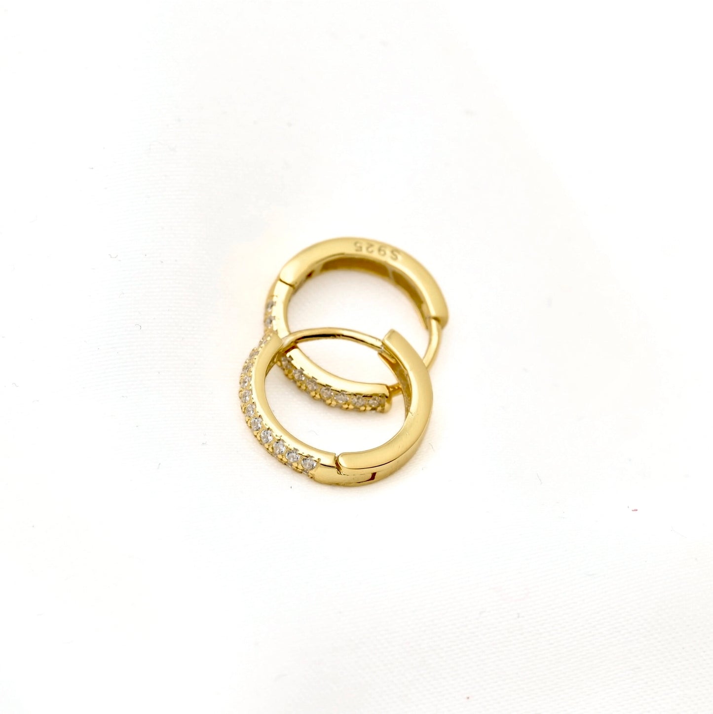 A pair of gold huggie hoop earrings, one plain and one paved with small clear stones, displayed on a white textured background.