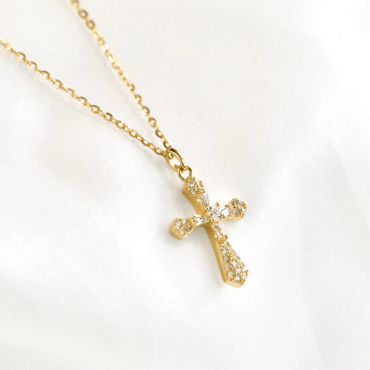 A gold cross pendant necklace adorned with small clear crystals, resting on a white satin fabric background.