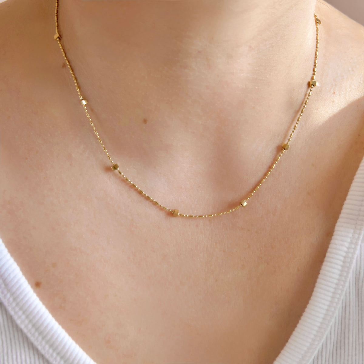 A delicate gold chain necklace with small spaced beads, worn on a person's neck over a white ribbed top.