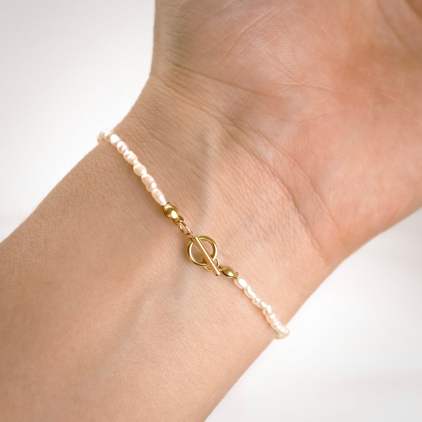 A delicate bracelet with small white pearls and gold-toned toggle clasp details, shown worn on a person's wrist against a soft white background.