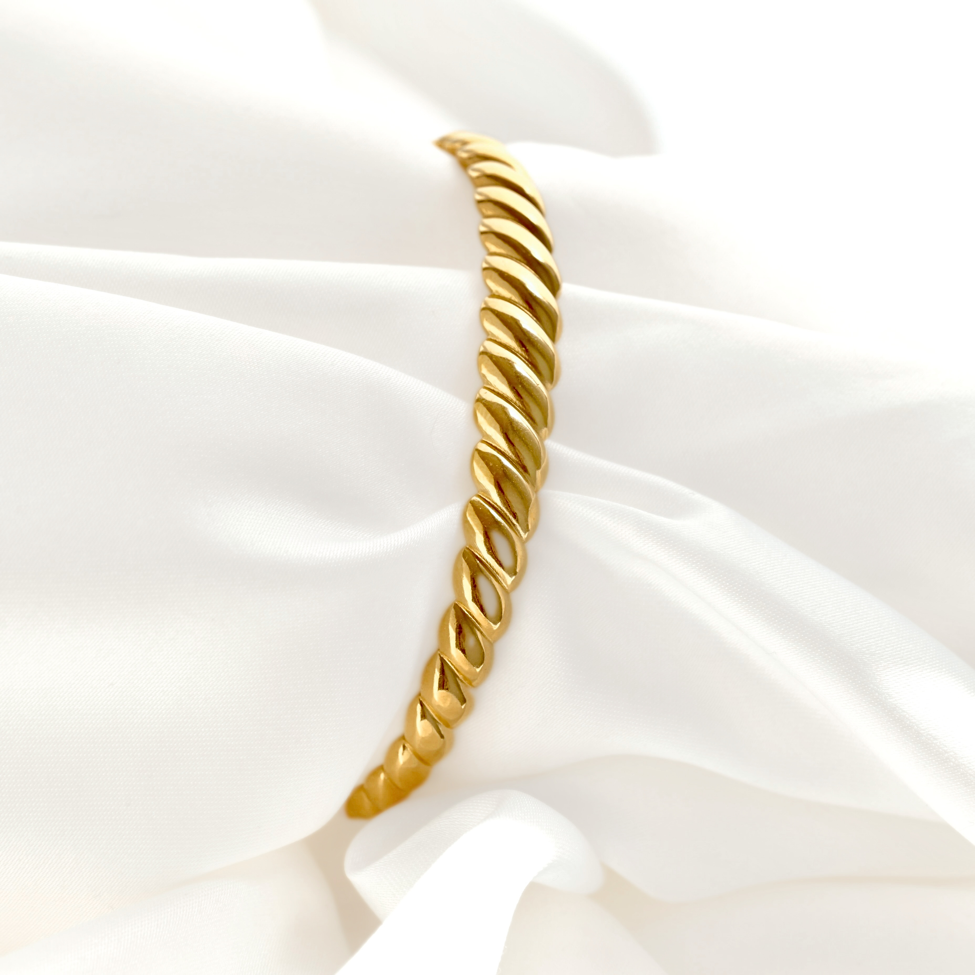 A gold twisted rope bangle bracelet displayed on a draped white satin fabric background.