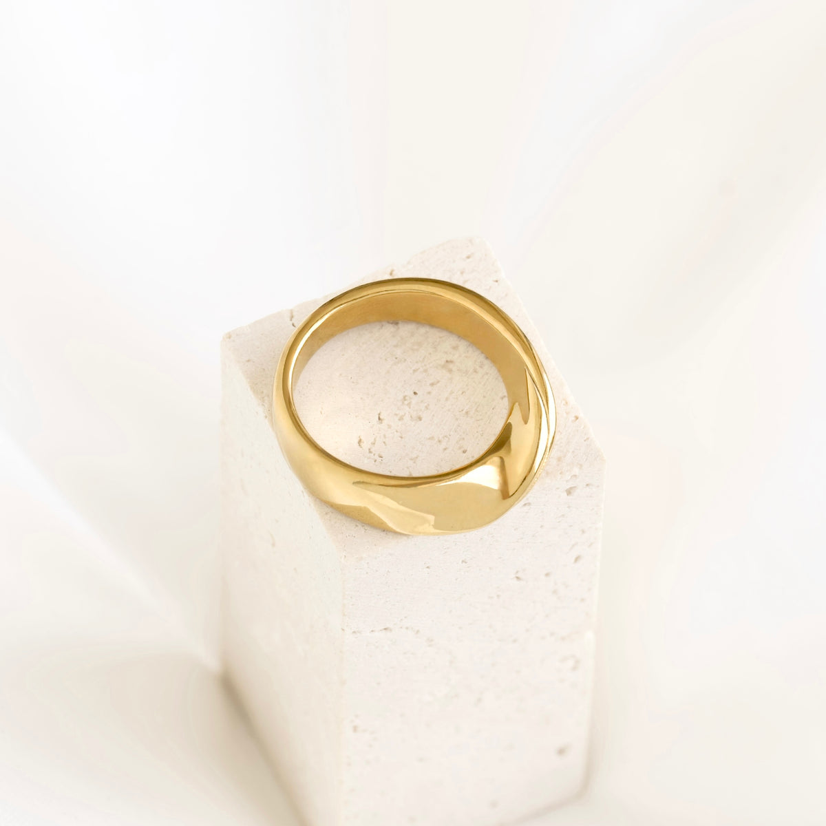 A polished gold ring displayed on a textured white stone block against a soft white background.