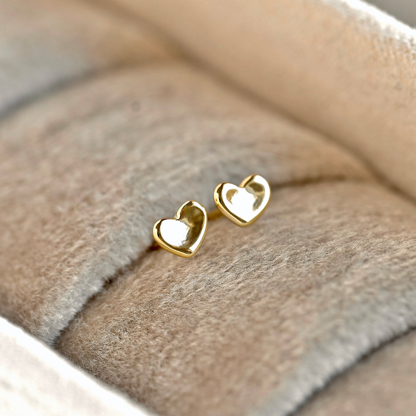 A pair of tiny gold heart studs resting on a soft beige velvet jewelry display.