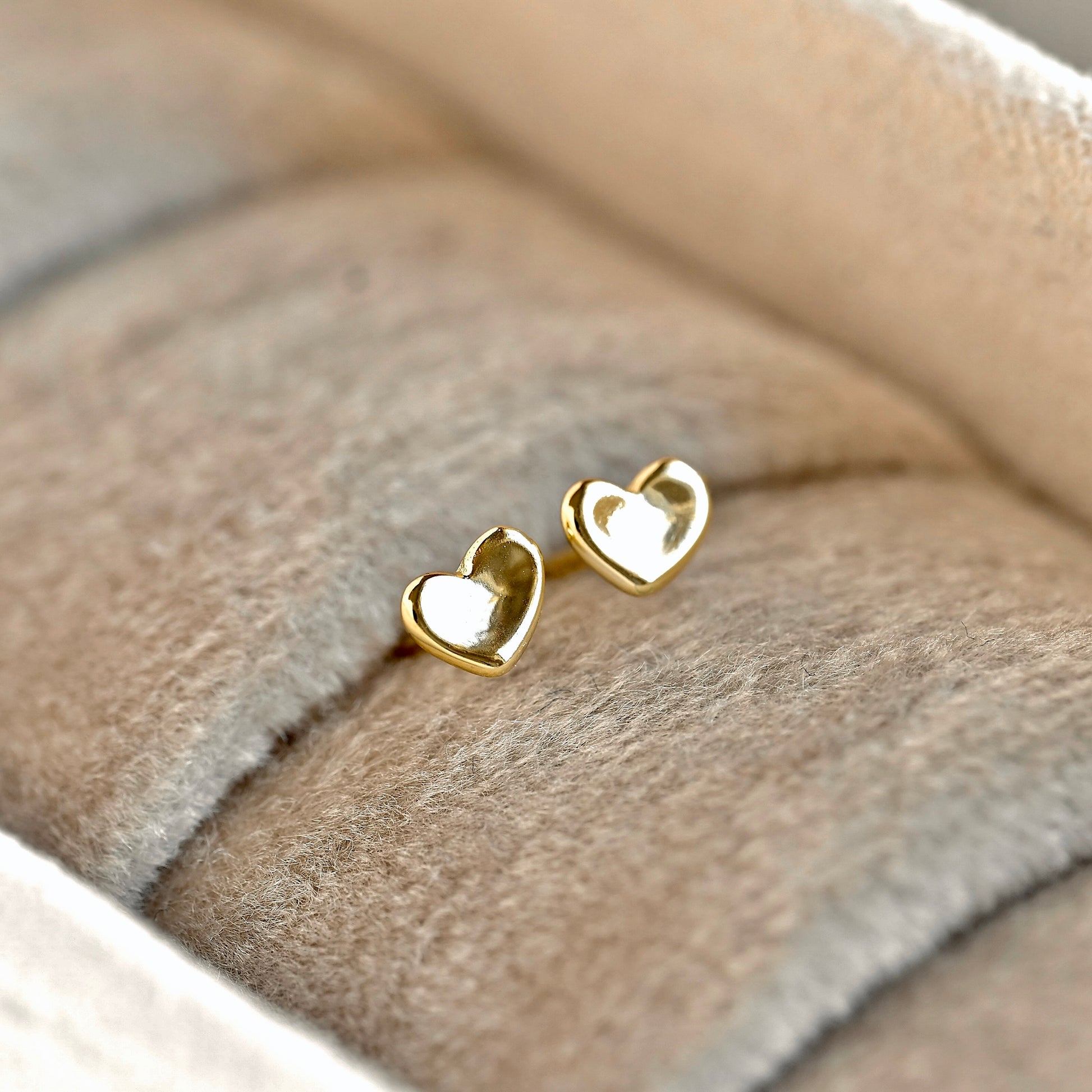 A pair of tiny gold heart studs resting on a soft beige velvet jewelry display.