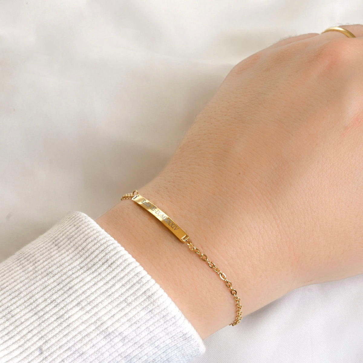 A delicate gold bar bracelet with engraved text, worn on a person's wrist over a white ribbed cuff, shown against a soft white fabric background.