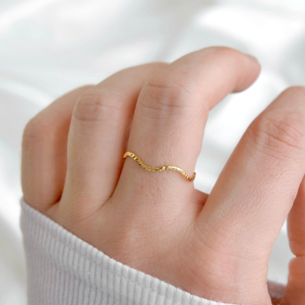 A delicate gold wavy ring worn on a person's ring finger, shown in a close-up shot against a soft white fabric background.