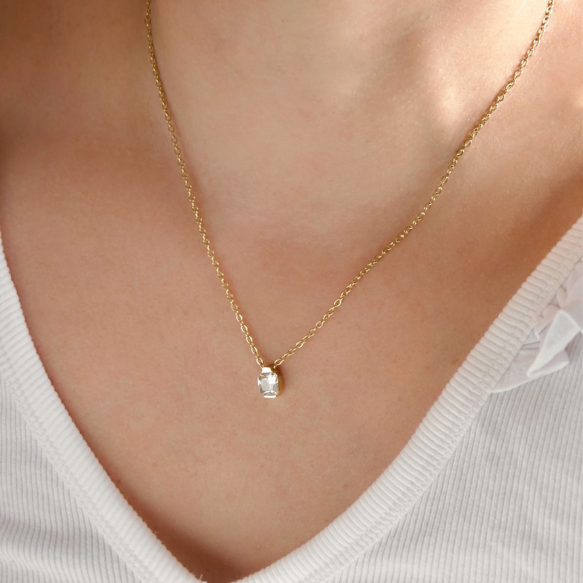 A delicate gold chain necklace with a small clear rectangular gemstone pendant, worn on a model's neck over a white ribbed top.