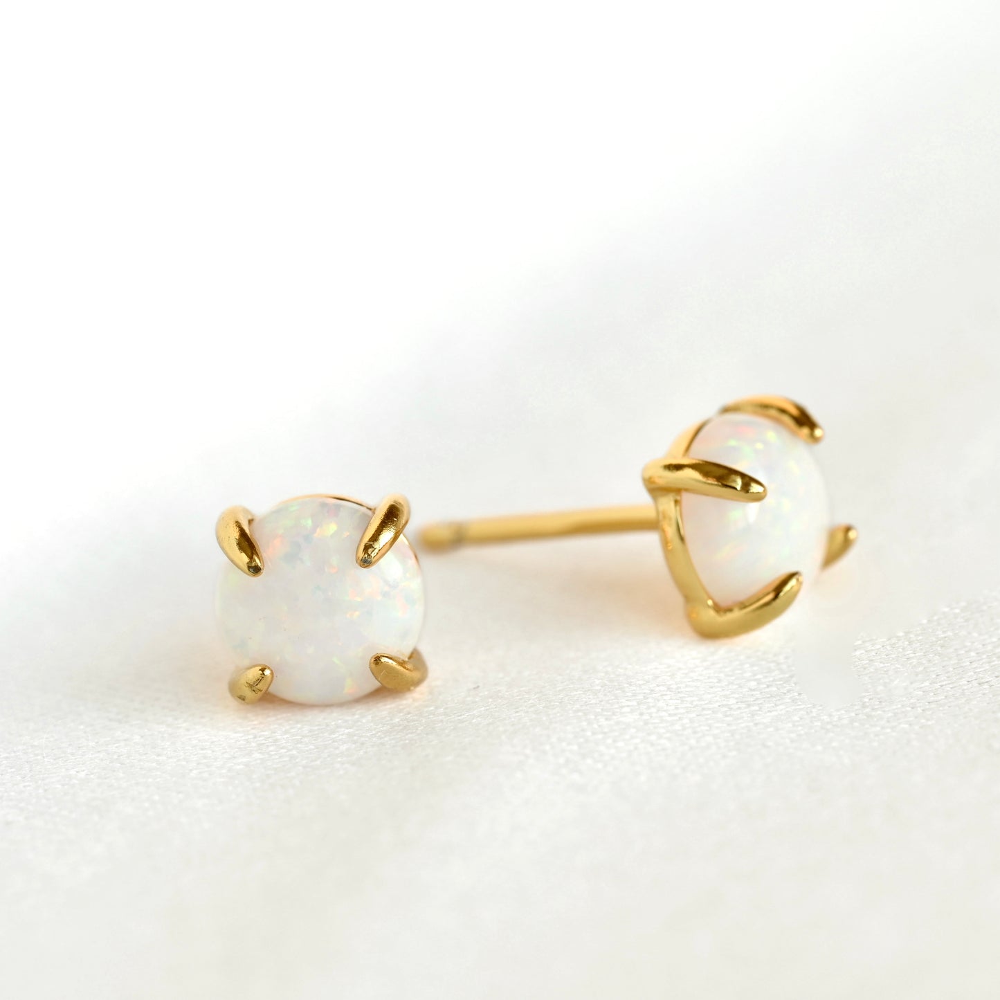 A pair of gold stud earrings with oval white opal gemstones in a four-prong setting, displayed on a clean white textured surface.