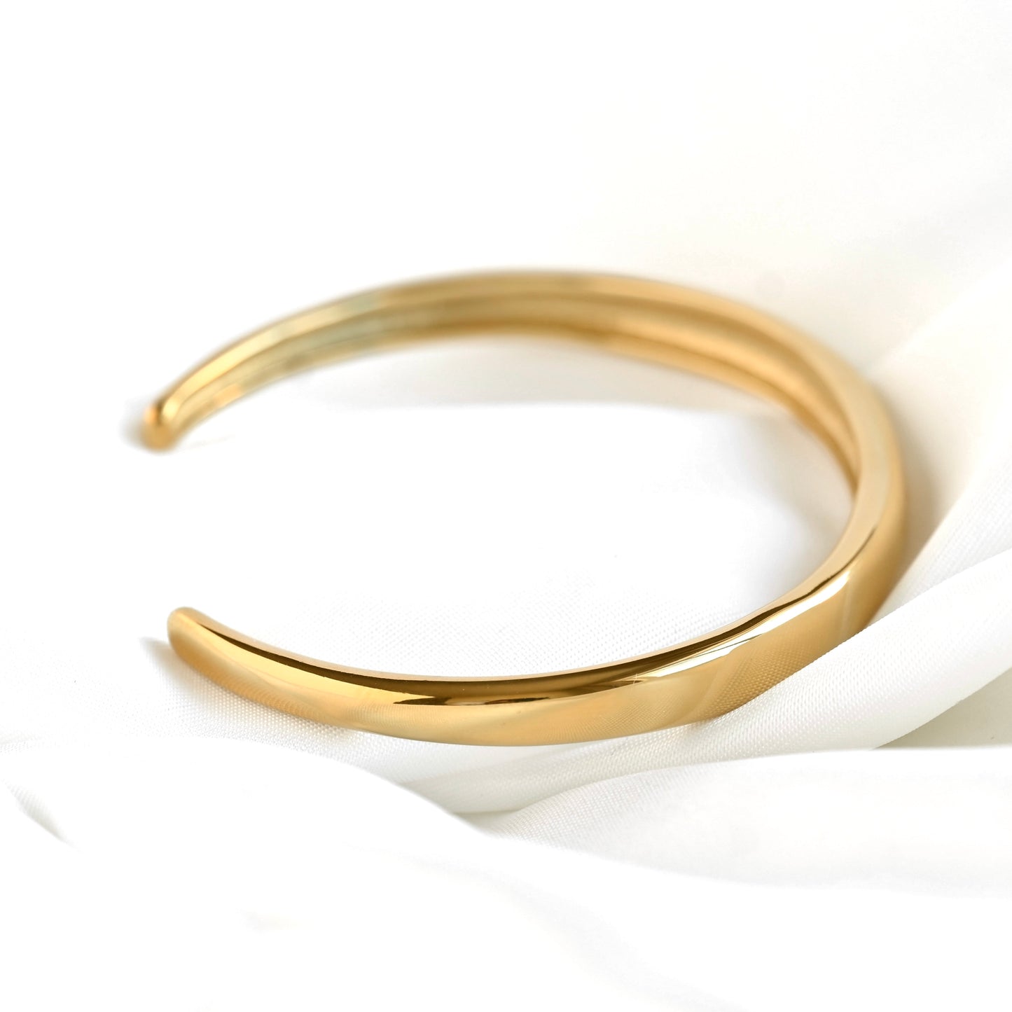 A polished gold-tone open cuff bracelet resting on draped white fabric.