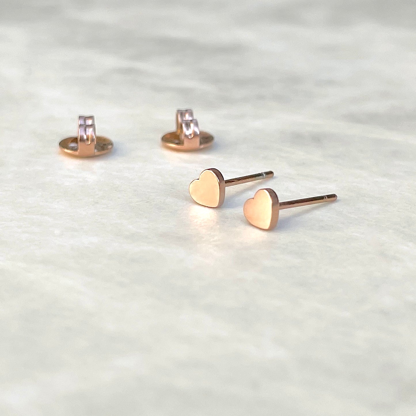 Angled front view of rose gold heart stud earrings with their butterfly backs  at the back on a light gray background