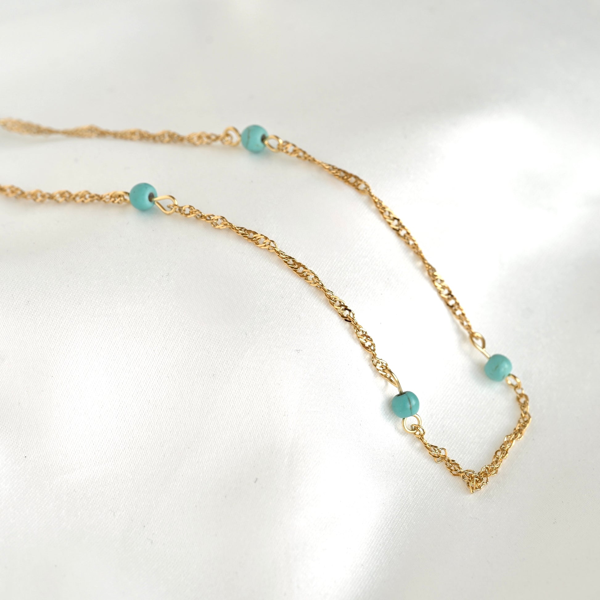 A delicate gold chain necklace with four small, round turquoise blue beads spaced along the chain, displayed on a soft white fabric background.
