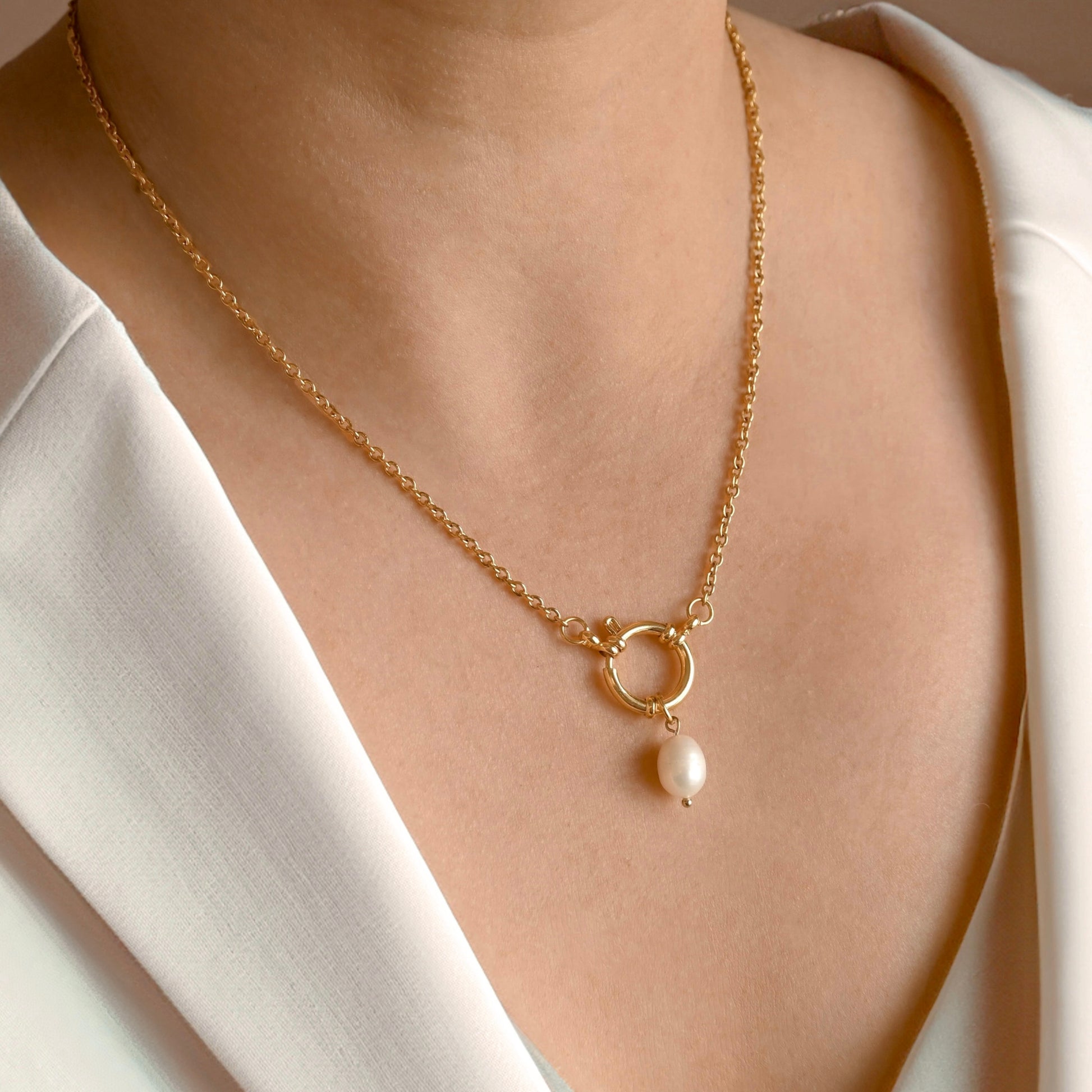 A close-up of a gold chain necklace with a sailor-clasp pendant and a dangling white pearl, worn by a model in a white blouse.