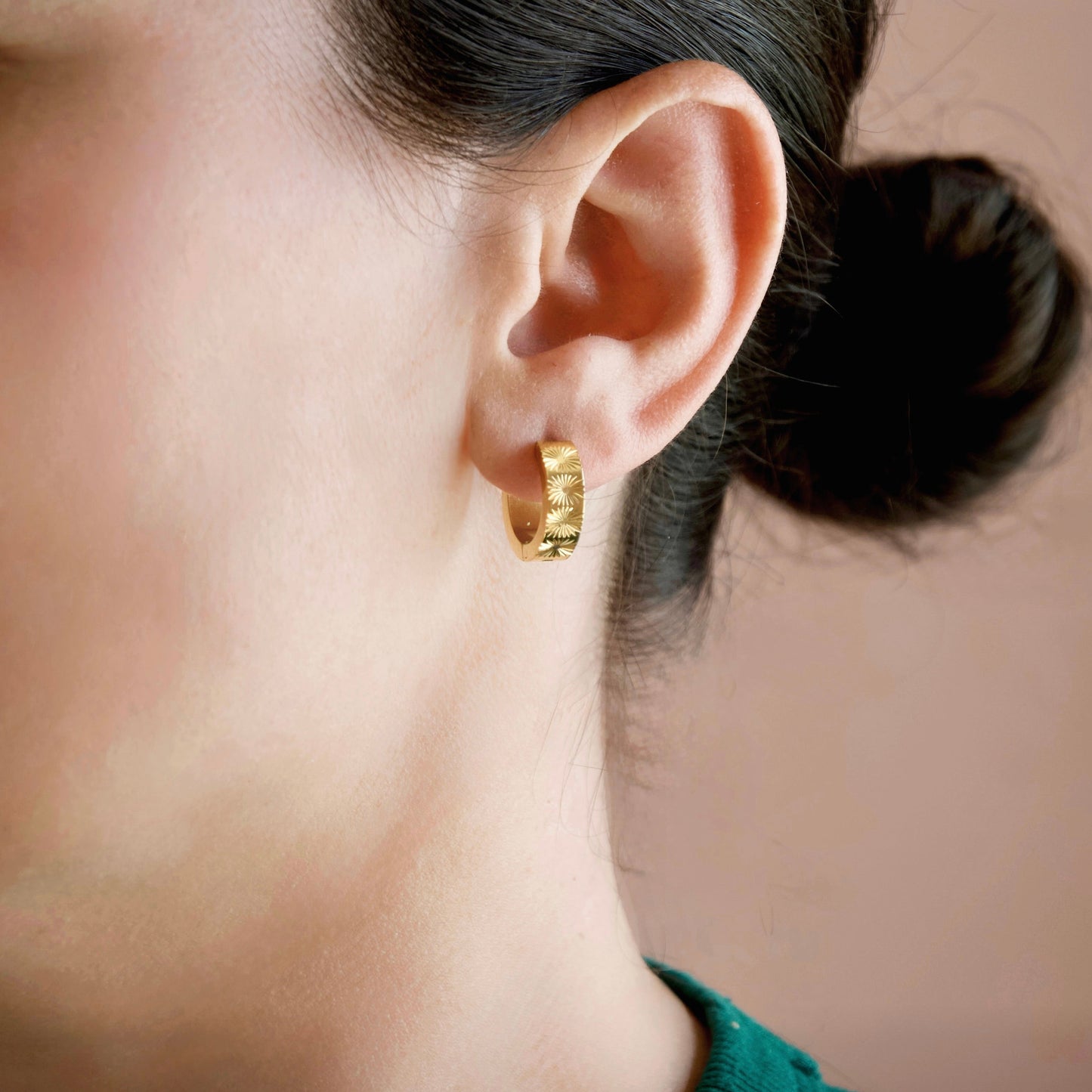 Person with black hair in a lower bun wearing gold sunburst hoop earrings.