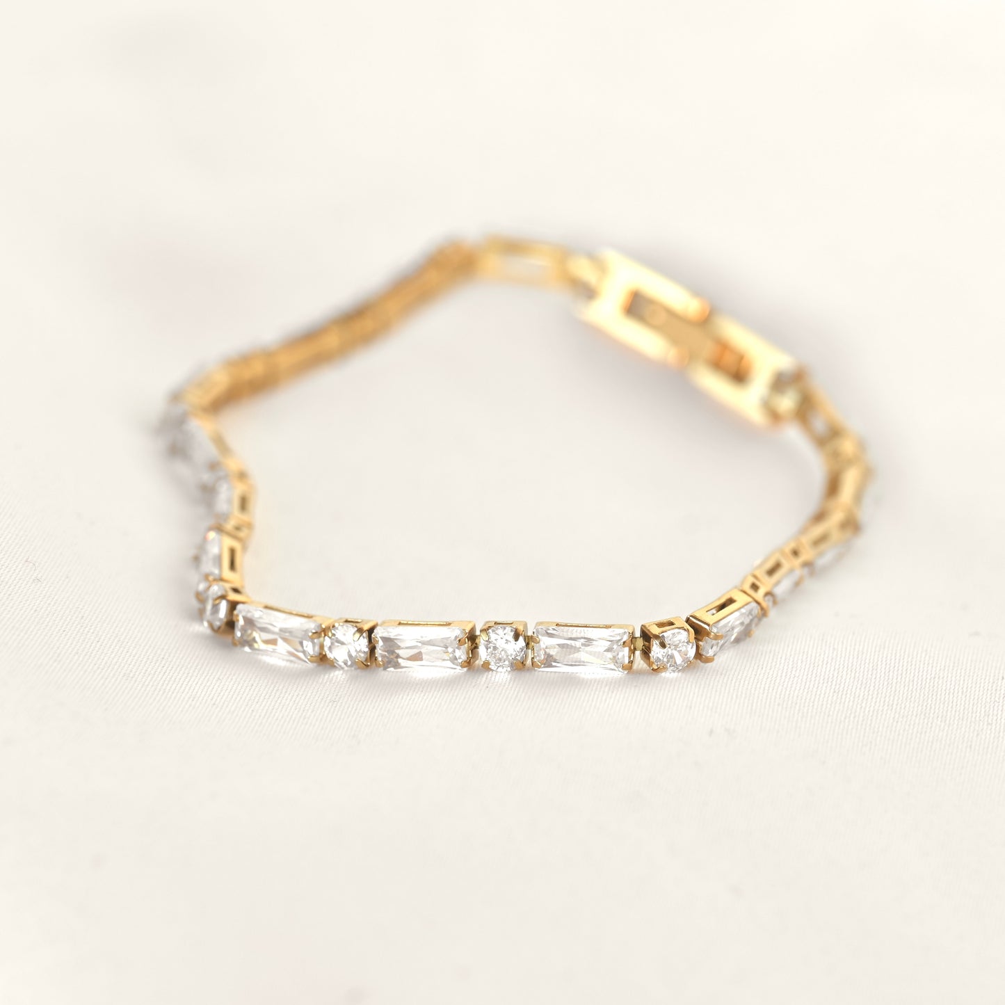 A yellow gold tennis bracelet featuring baguette and round cut clear crystals, displayed on a soft, light beige fabric background.
