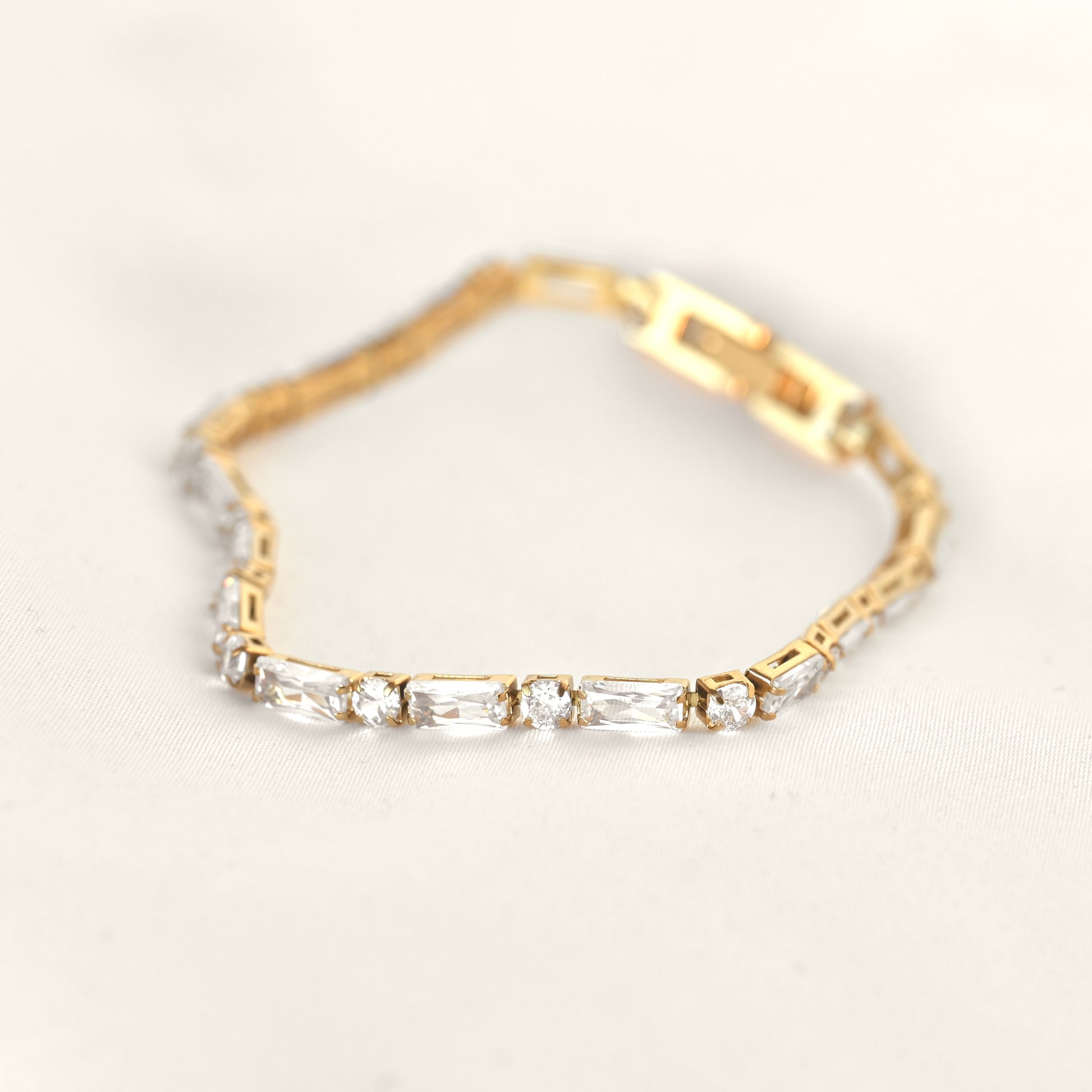 A yellow gold tennis bracelet featuring baguette and round cut clear crystals, displayed on a soft, light beige fabric background.