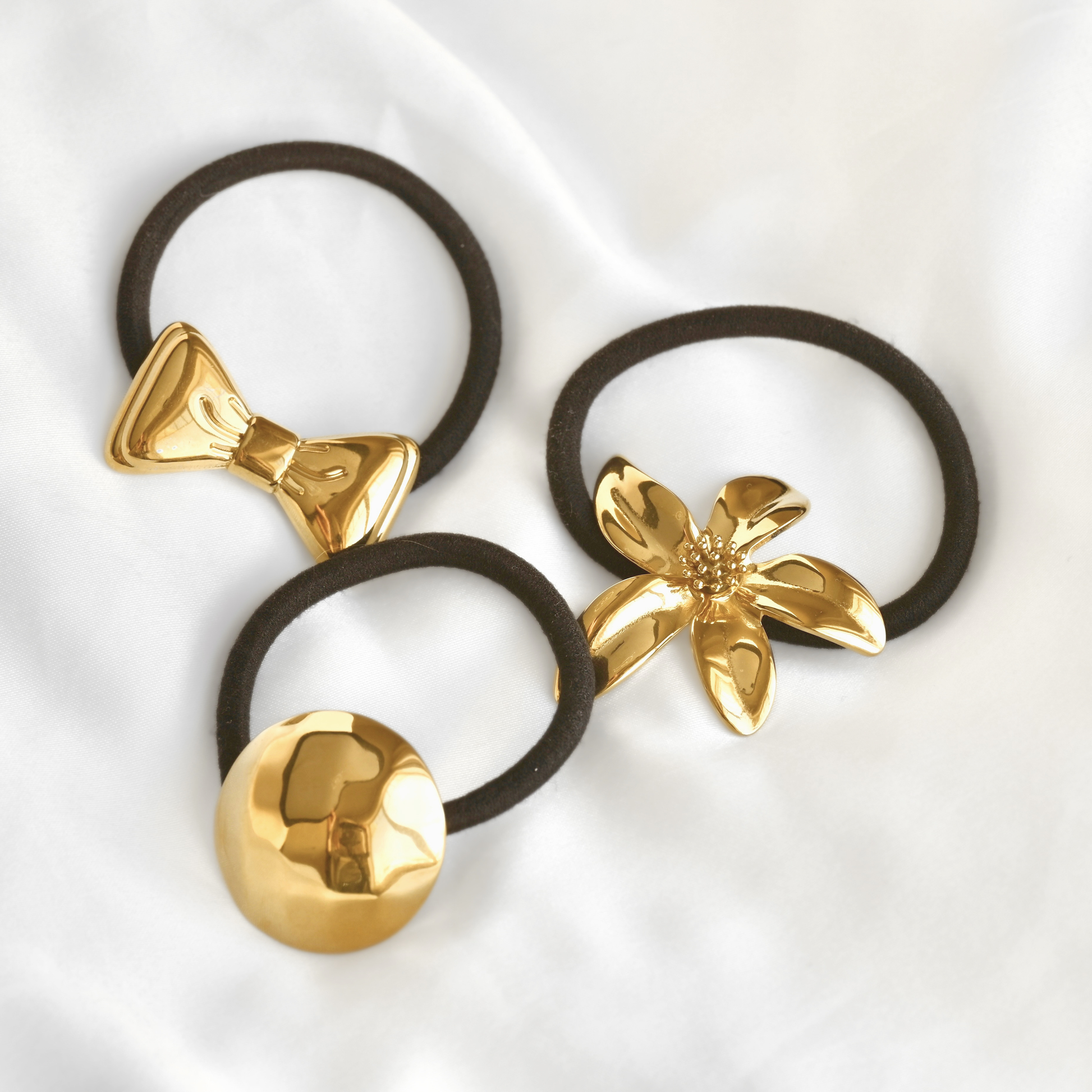 Three black elastic hair ties with shiny gold decorative accents, including a bow, a flower, and a round disc, arranged on a white fabric background.