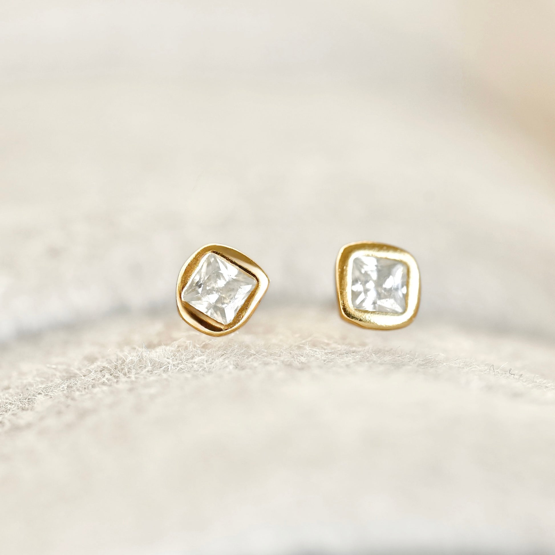 A pair of gold stud earrings with square cushion-cut clear gemstones, resting on a soft, textured beige fabric surface.