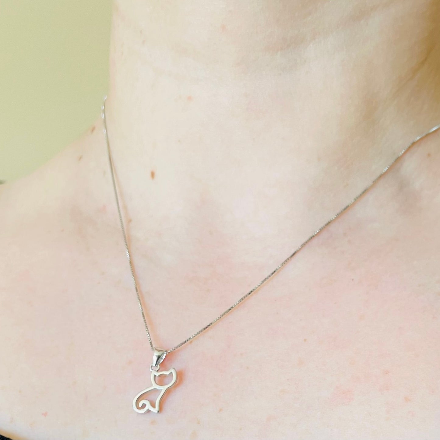 Silver necklace with sitting cat silhouette pendant on a person's neck against a neutral background