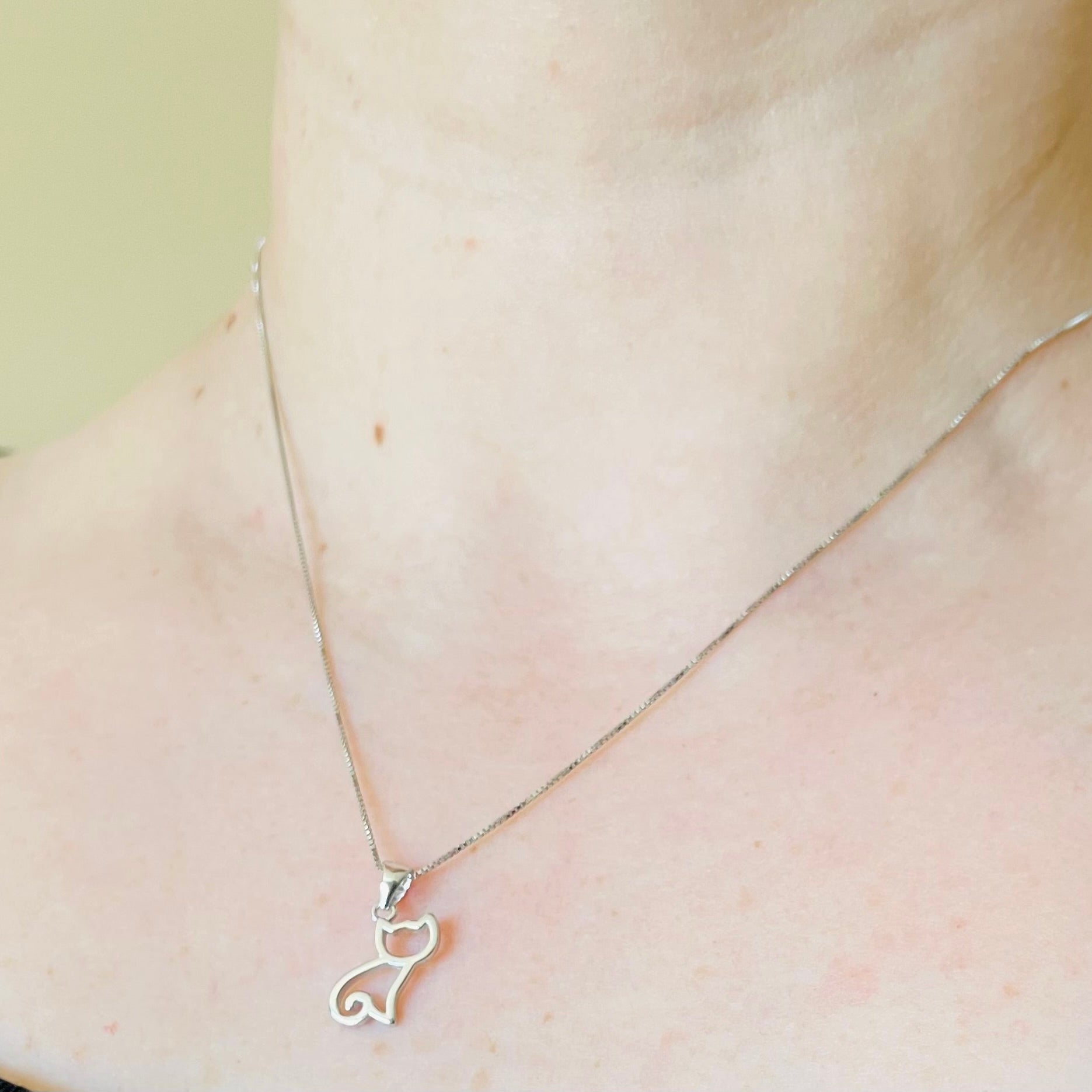 Silver necklace with sitting cat silhouette pendant on a person's neck against a neutral background