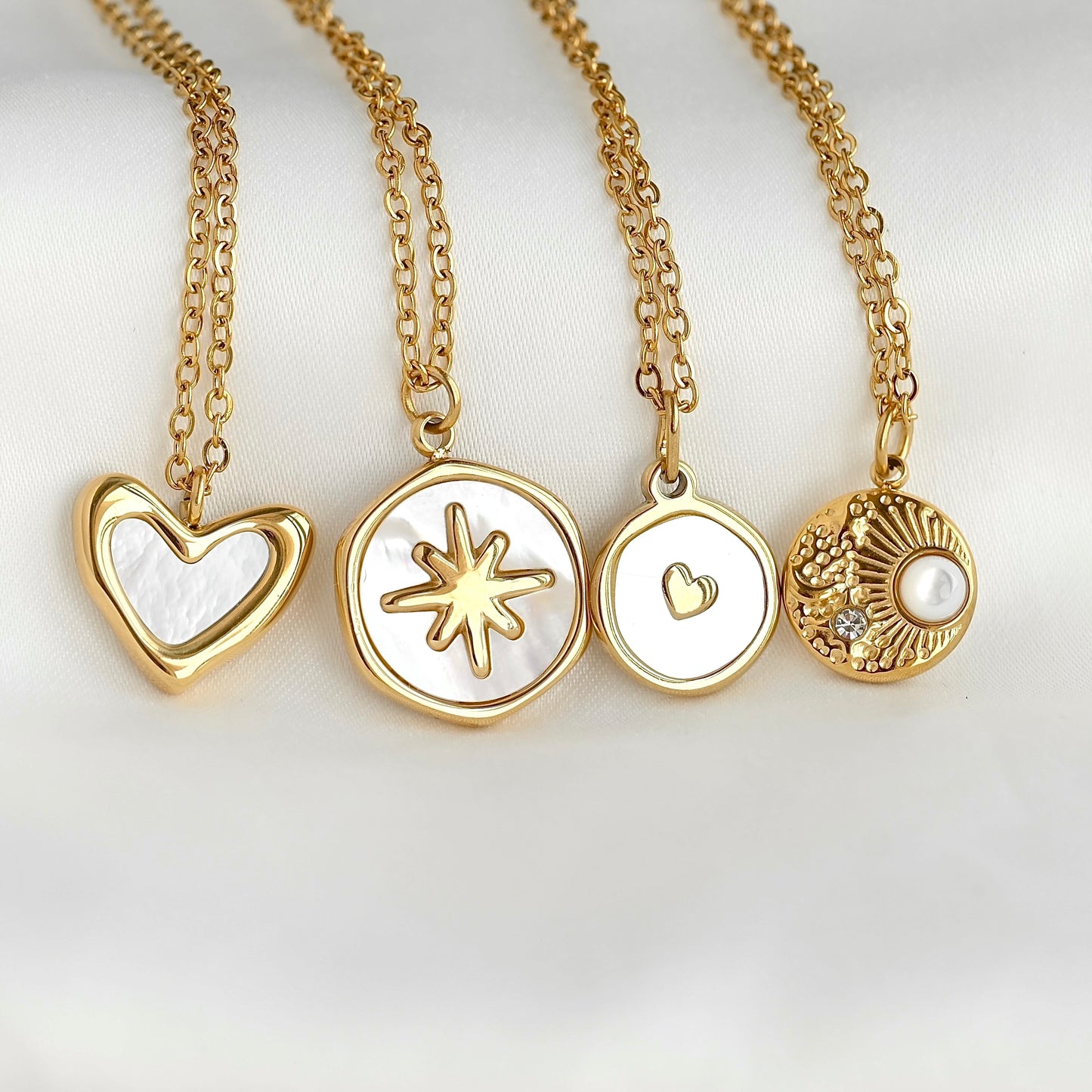 Four gold pendant necklaces with white inlay details, displayed on a soft white background. The pendants include a heart, a starburst, a small heart, and a textured sunburst with a pearl.