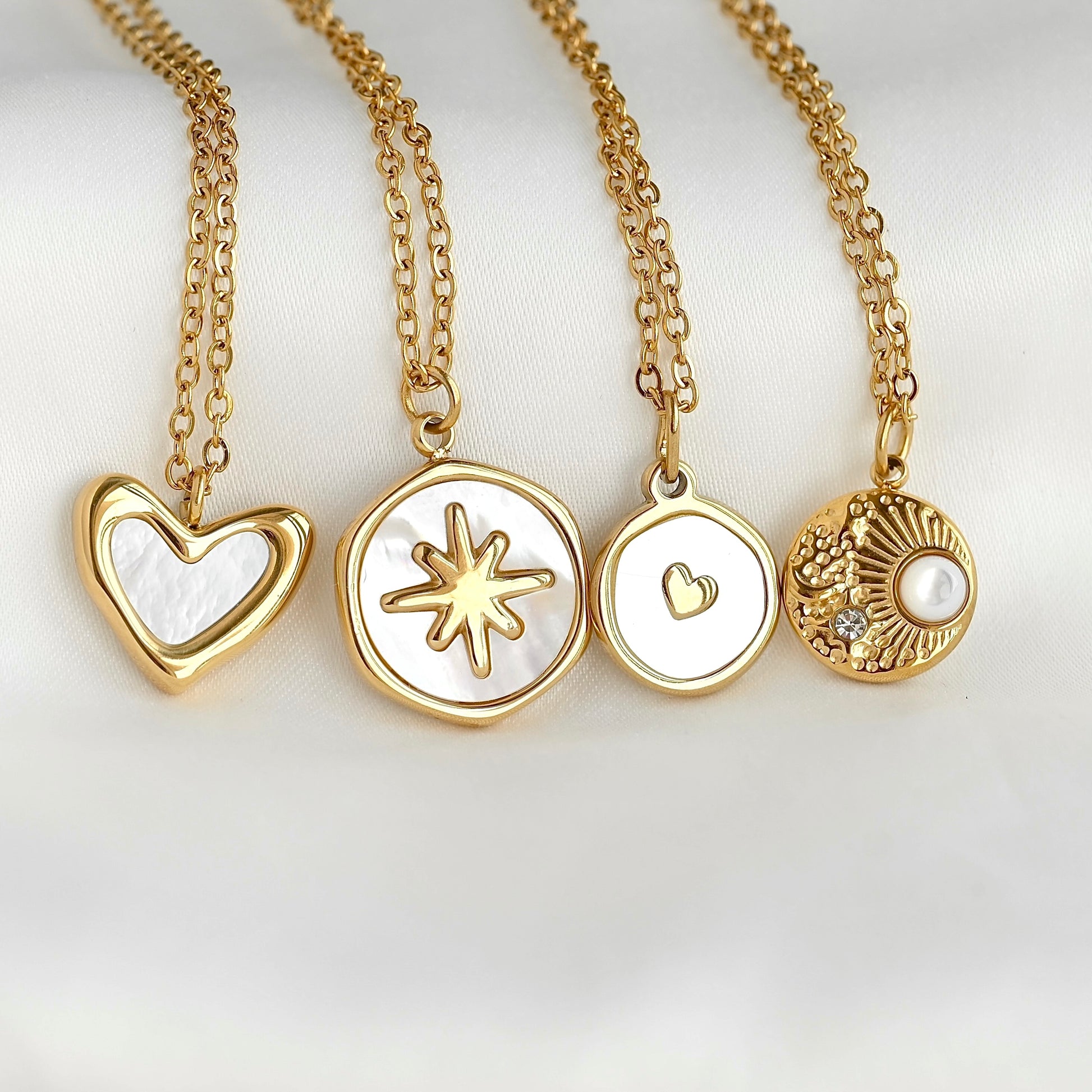 Four gold pendant necklaces with white inlay details, displayed on a soft white background. The pendants include a heart, a starburst, a small heart, and a textured sunburst with a pearl.