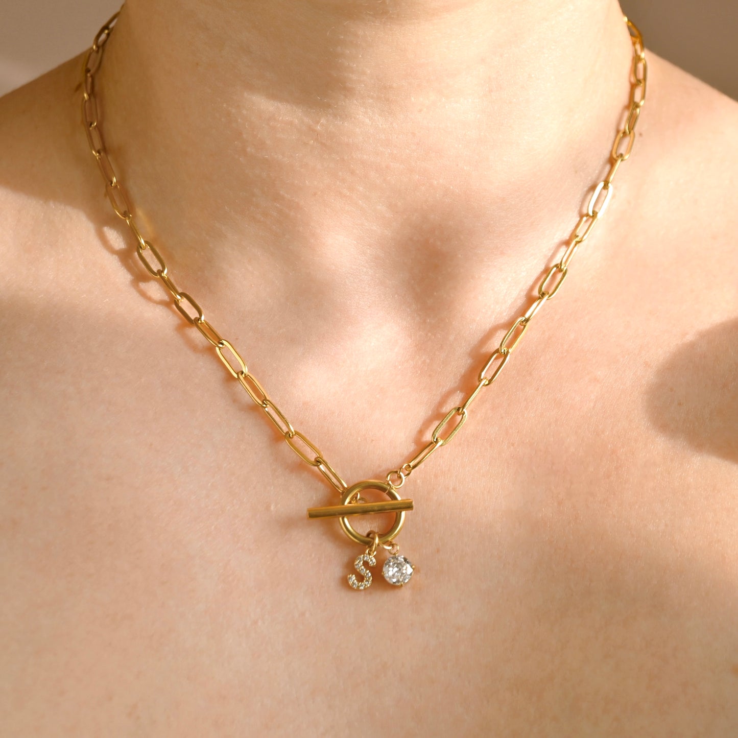 A gold paperclip chain necklace with a toggle clasp and two small dangling clear initial and april birthstone, shown worn on a person's neck and upper chest.