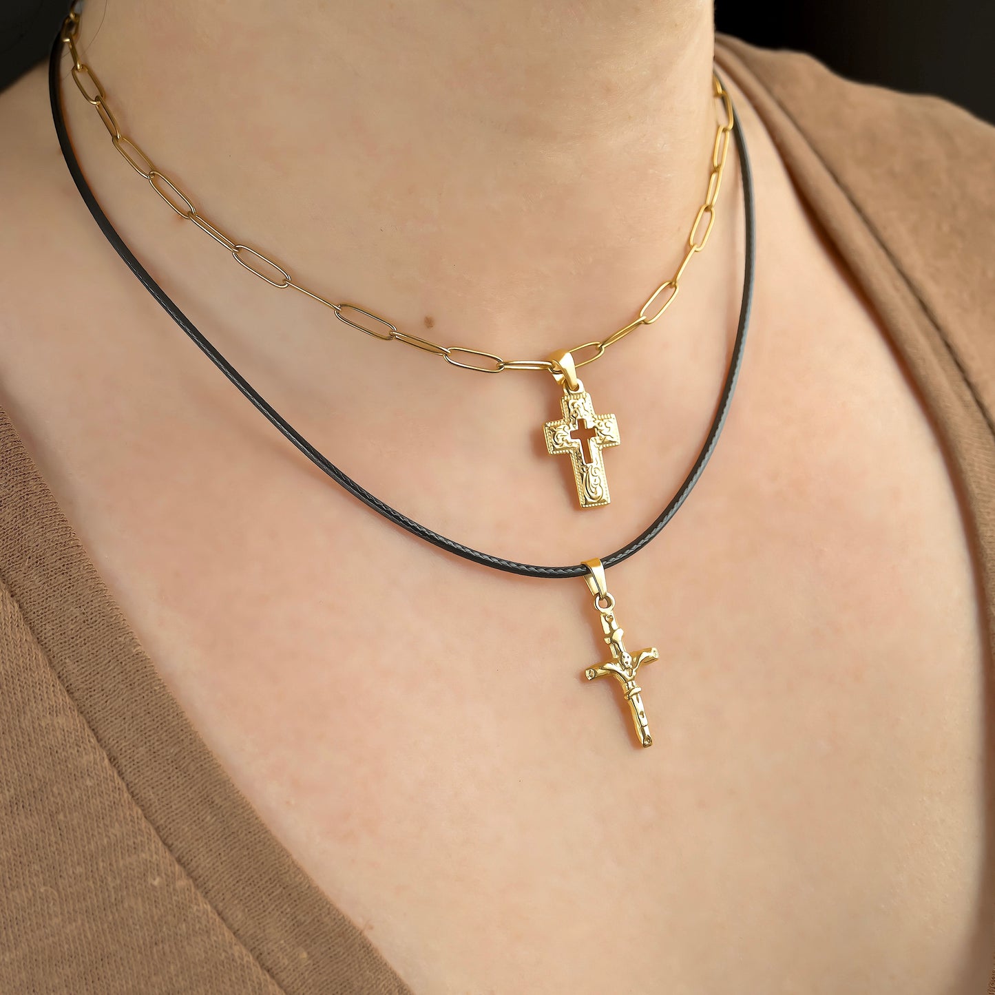 Two gold cross necklaces, one with a thick pendant on a paperclip chain, the other cross with Jesus pendant on a black rope chain, on a person wearing a brown top.