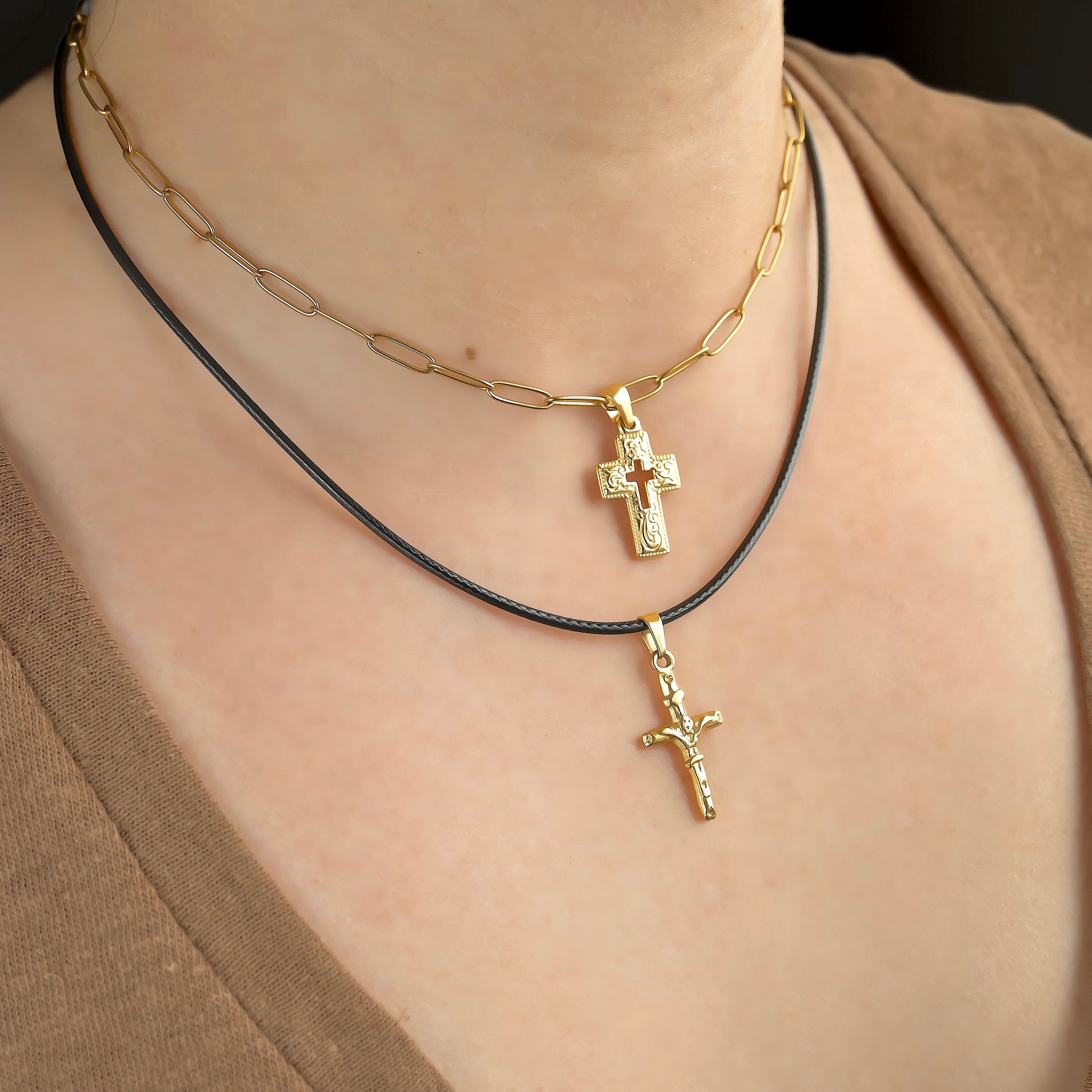 Two gold cross necklaces, one with a thick pendant on a paperclip chain, the other cross with Jesus pendant on a black rope chain, on a person wearing a brown top.