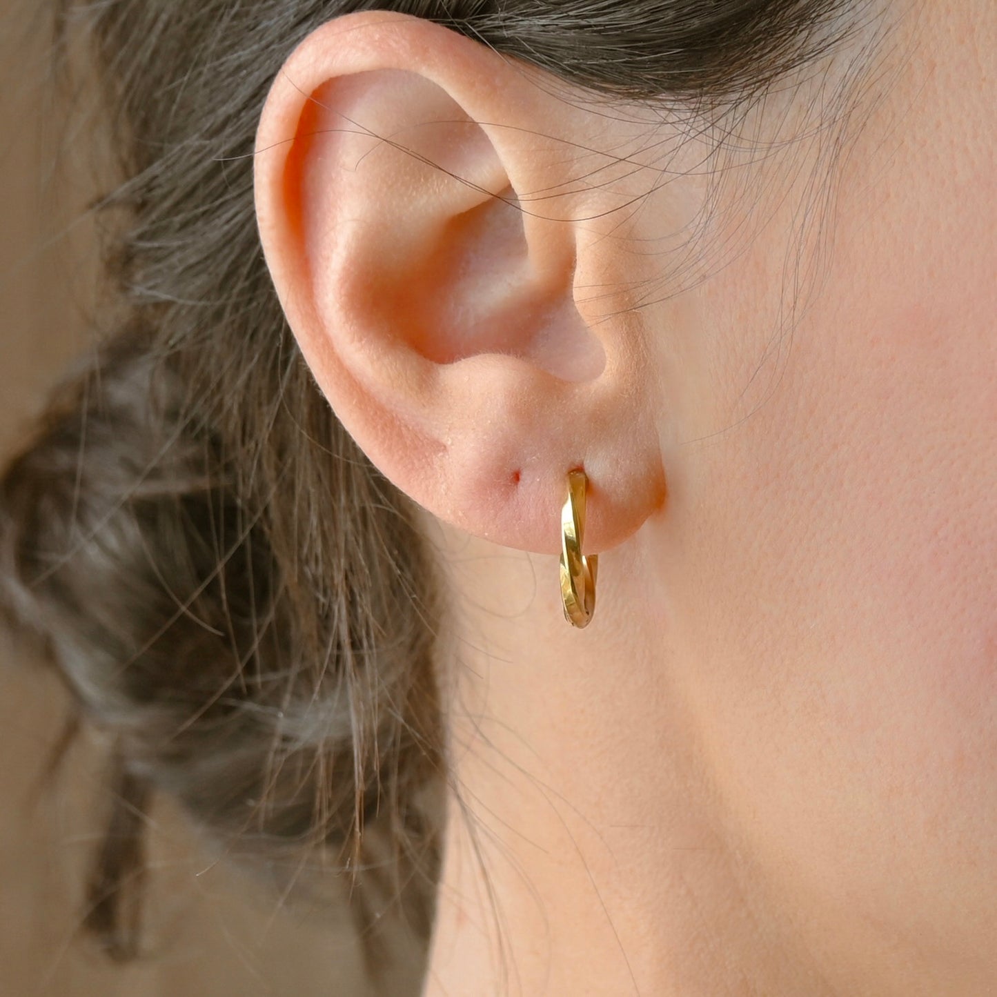 A close-up of a person's ear wearing a small twisted gold hoop earring.