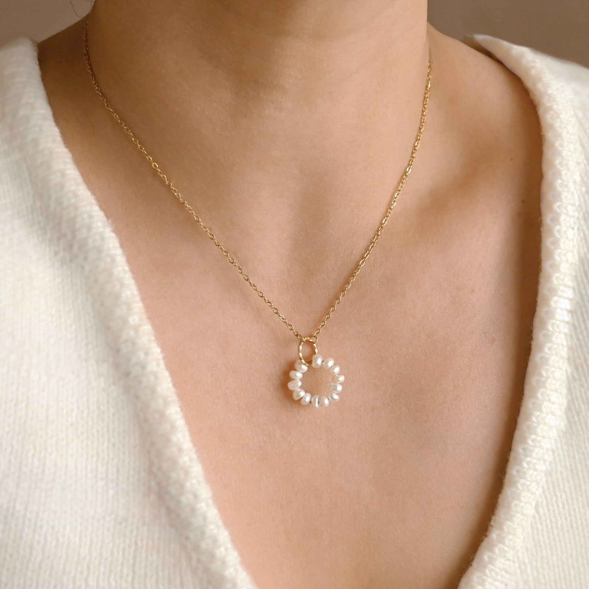 A close-up of a person wearing a delicate gold chain necklace with a small circular white pearl pendant suspended from an interlocking gold swirl circle, styled with a white knit V-neck top.