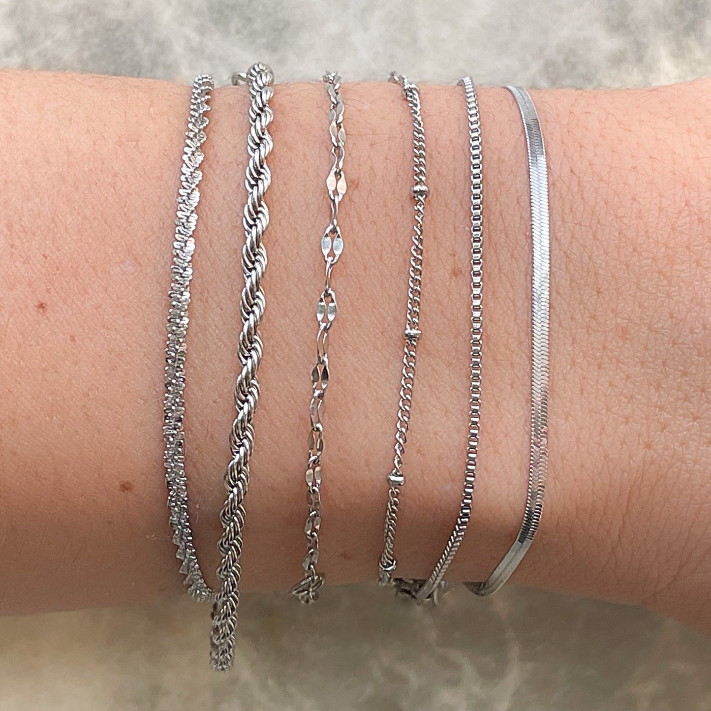 Close-up side view of silver chain bracelets. From left to right: sparkle rope, 2mm twisted tope, dainty mariner, satellite, box and thin flat snake chains on a person's wrist against a neutral background
