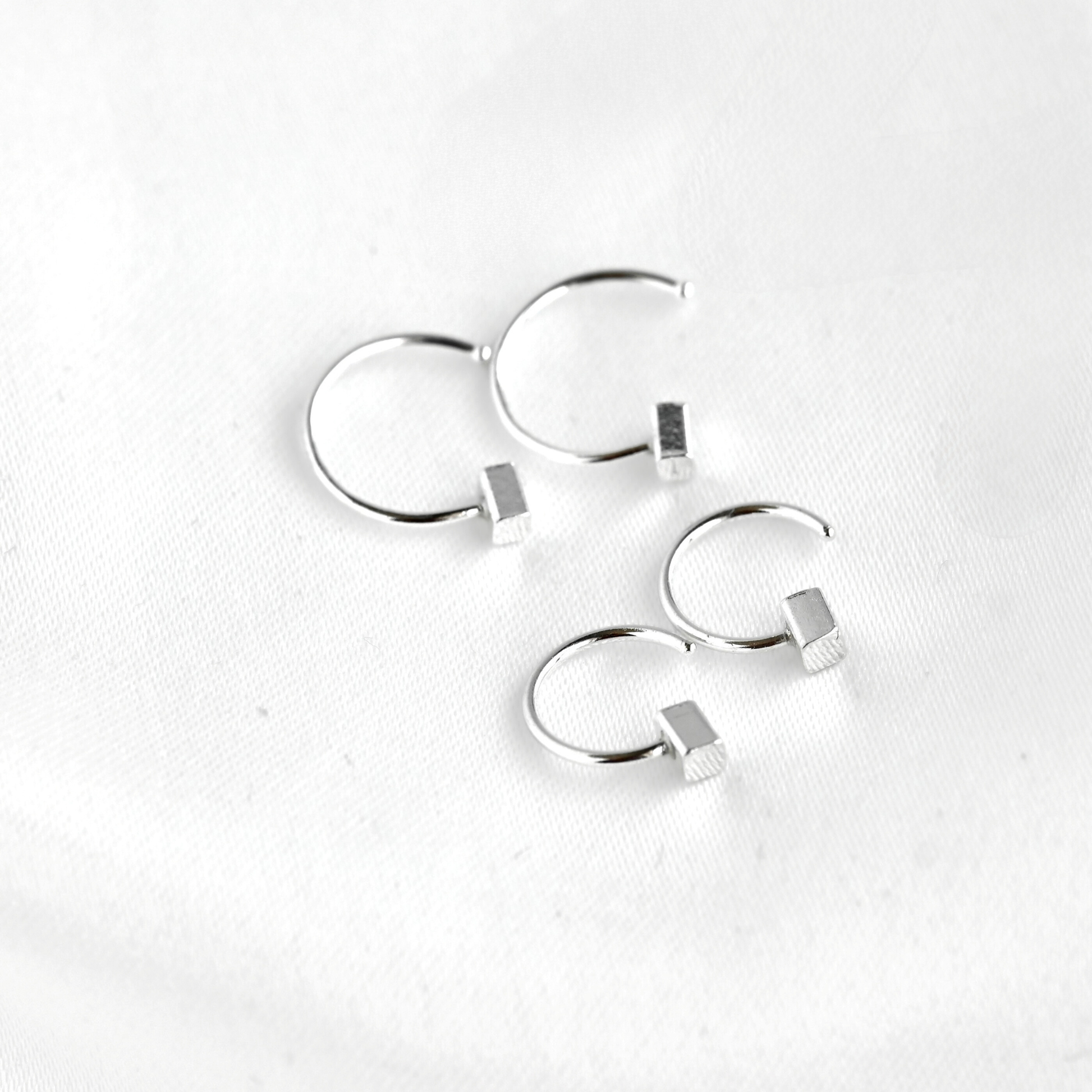 Four silver hoop earrings with small rectangular bead details, arranged on a white textured fabric background.