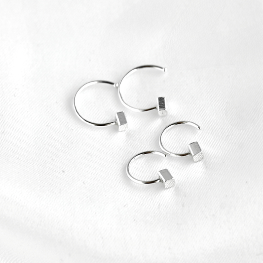 Four silver hoop earrings with small rectangular bead details, arranged on a white textured fabric background.