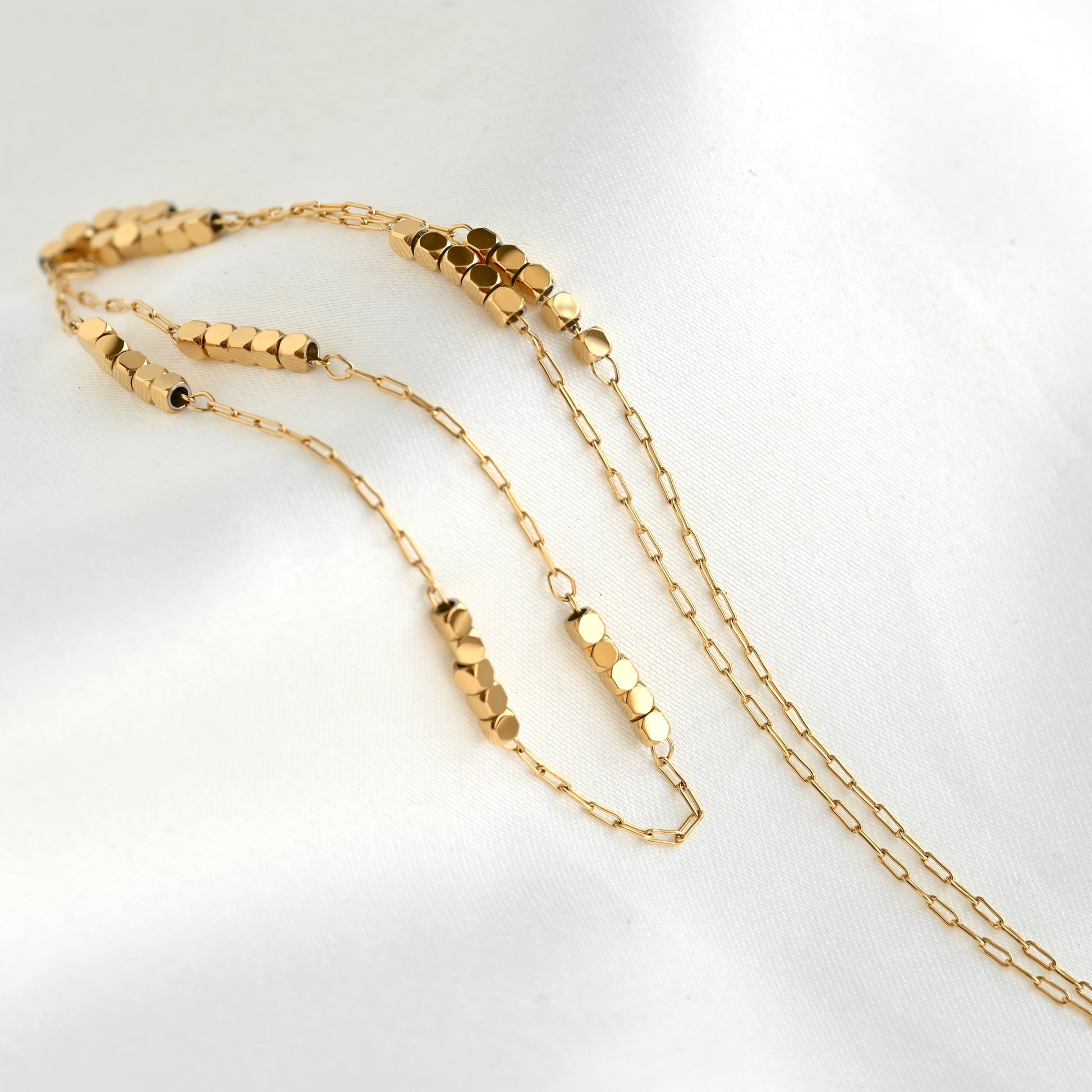 A gold chain necklace featuring textured, cube beads and an ultra thin paperclip chain design, displayed on a soft white fabric background.