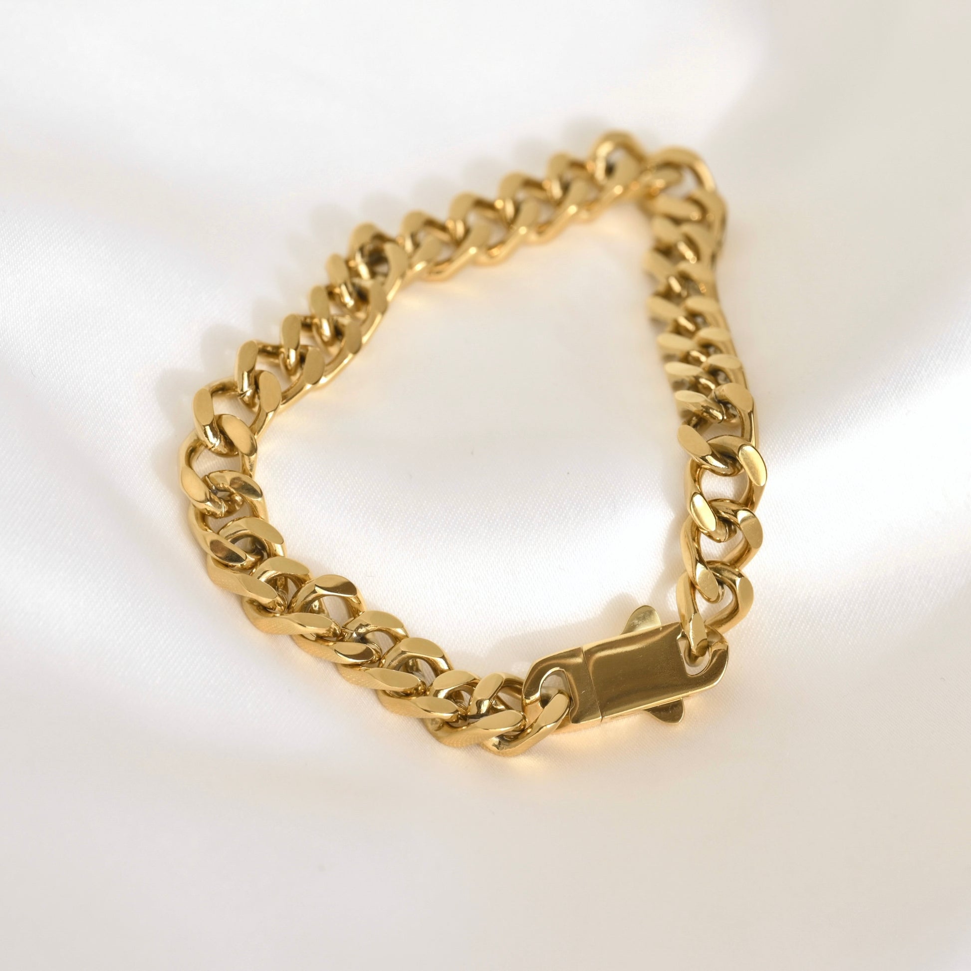 A gold-tone chunky curb link chain bracelet with a box clasp, displayed on a clean white satin fabric background.